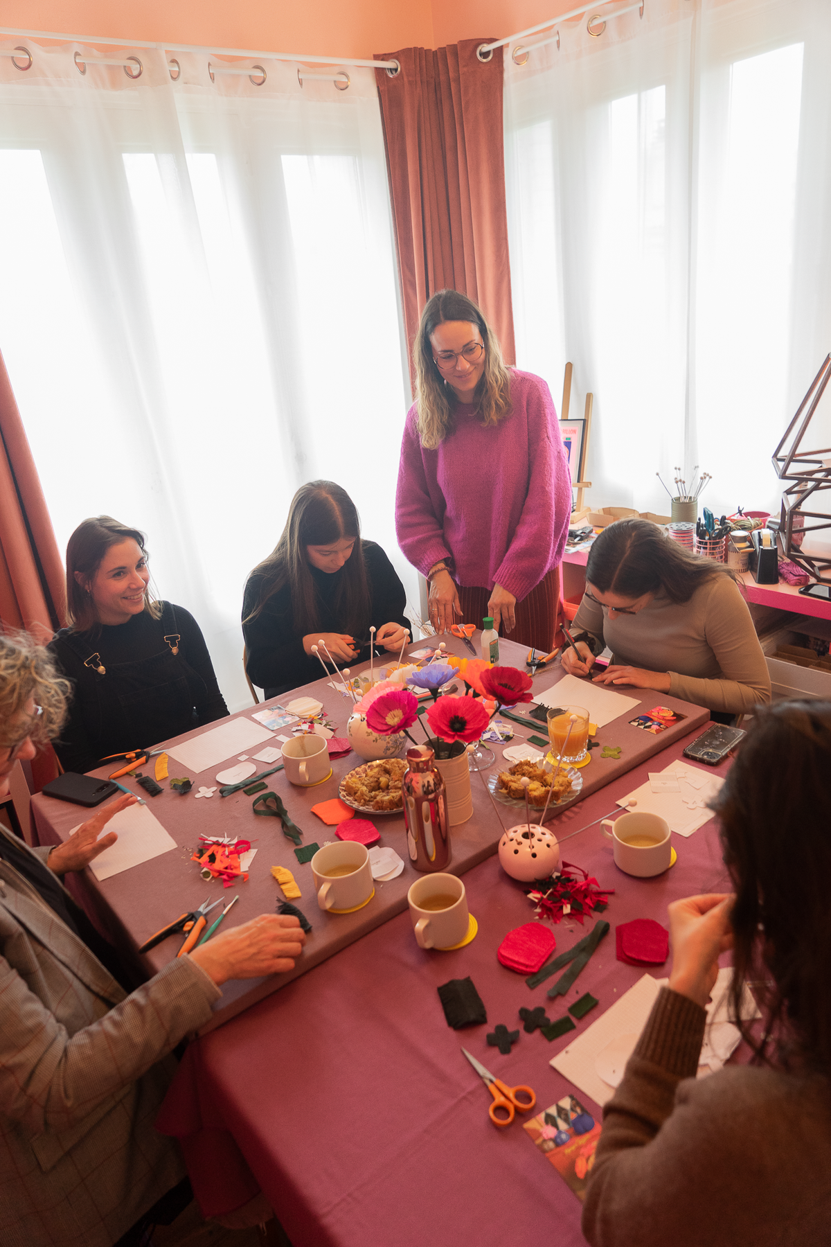 Vu d'ensemble d’un atelier fleurs en papier crépon avec 5 participantes autour d’une table guidées par l’animatrice en pull fushia avec au milieu de la table des fleurs, des snacks et des boissons, atelier créatif, team building, EVJF, animé par Maison Tessier.