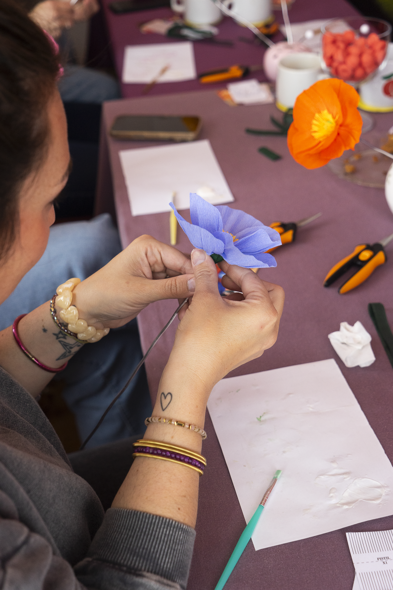 Vue plongeante sur les mains d’une participante assemblant des pétales pour créer un Pavot en papier crépon lors d’un atelier fleurs en papier, team building, EVJF, animé par Maison Tessier avec un Pavot orange en arrière-plan.