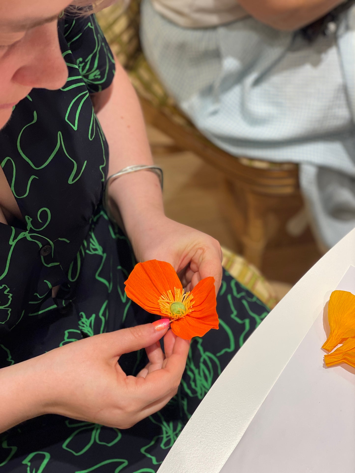 Vue plongeante sur les mains d’une participante en train d’assembler les pétales de sa fleur pour créer un Pavot en papier crépon orange lors d’un atelier fleurs en papier, team building, EVJF, animé par Maison Tessier.