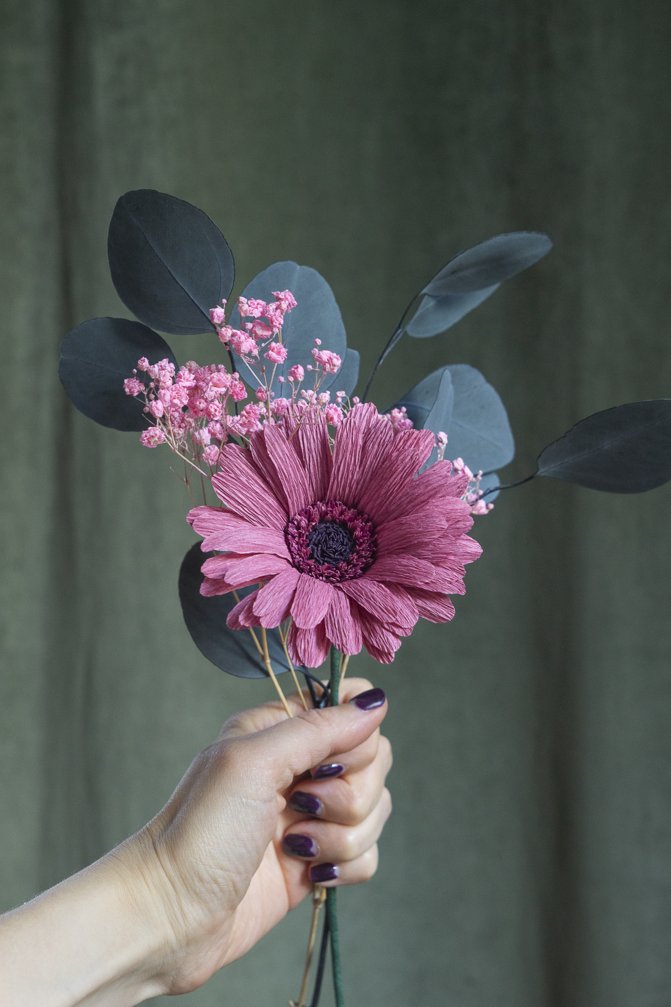 Bouquet d’un Gerbera vieux rose en papier crépon, accompagné de gypsophile rose et de feuilles d'eucalyptus séchées, tenus à la main sur fond vert kaki, fleurs éternelles artisanales faites main par Maison Tessier.