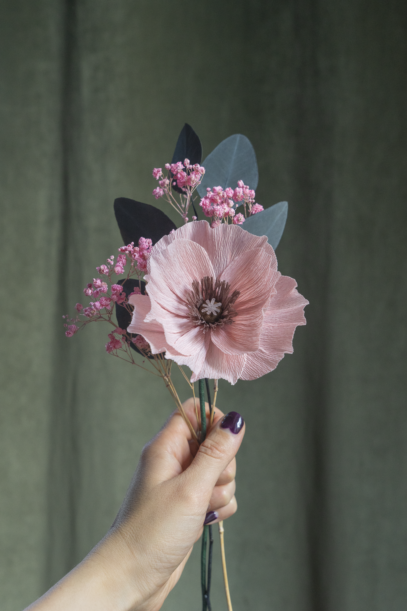 Bouquet d’un Pavot vieux rose clair en papier crépon, accompagné de gypsophile rose et de feuilles d'eucalyptus séchées, tenus à la main sur fond vert, fleurs éternelles artisanales faites main par Maison Tessier.
