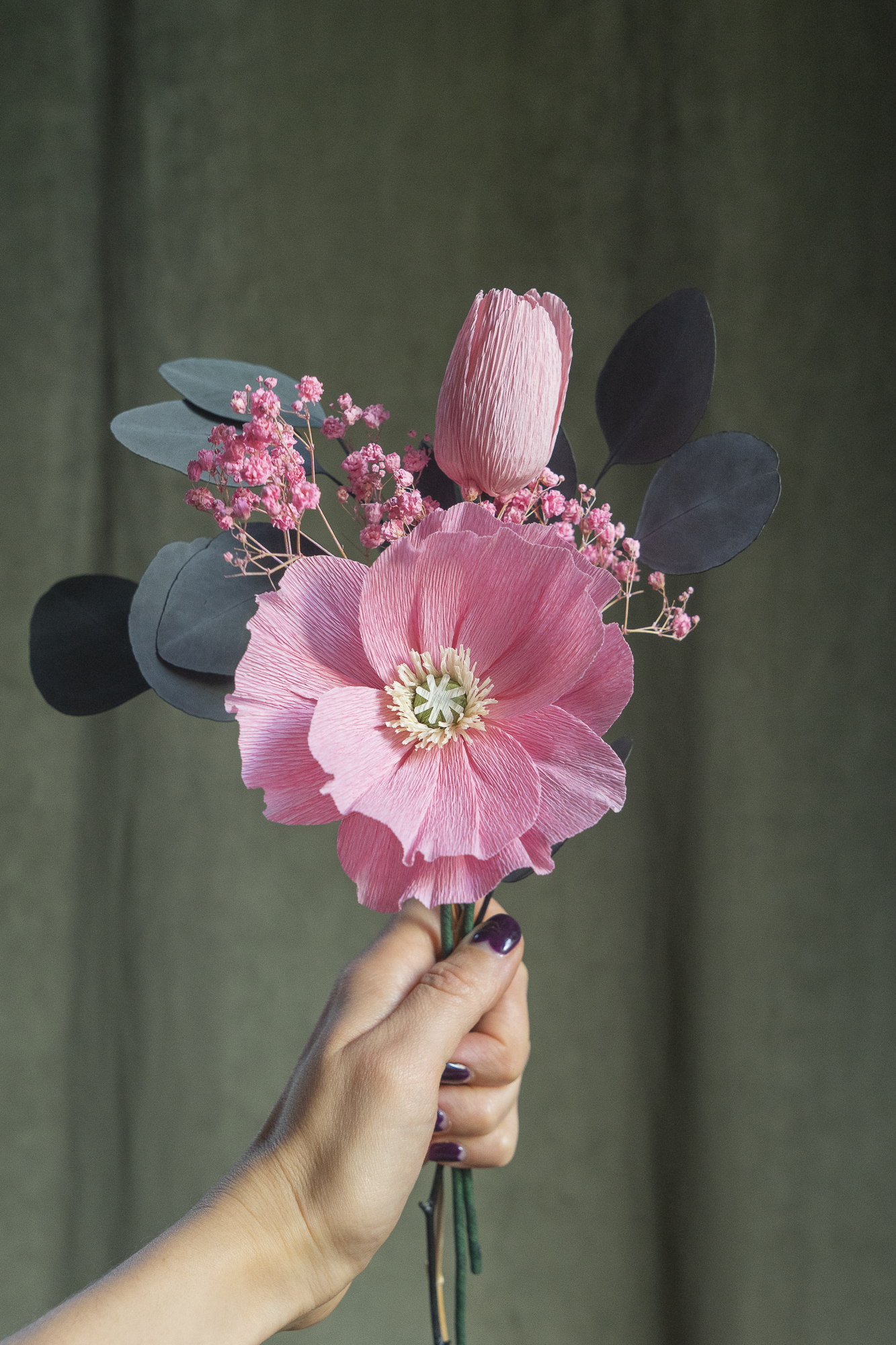 Bouquet d’un Pavot rose clair et d’une Tulipe rose clair en papier crépon, accompagné de gypsophile rose et de feuilles d'eucalyptus séchées, tenus à la main sur fond vert kaki, fleurs éternelles artisanales faites main par Maison Tessier.