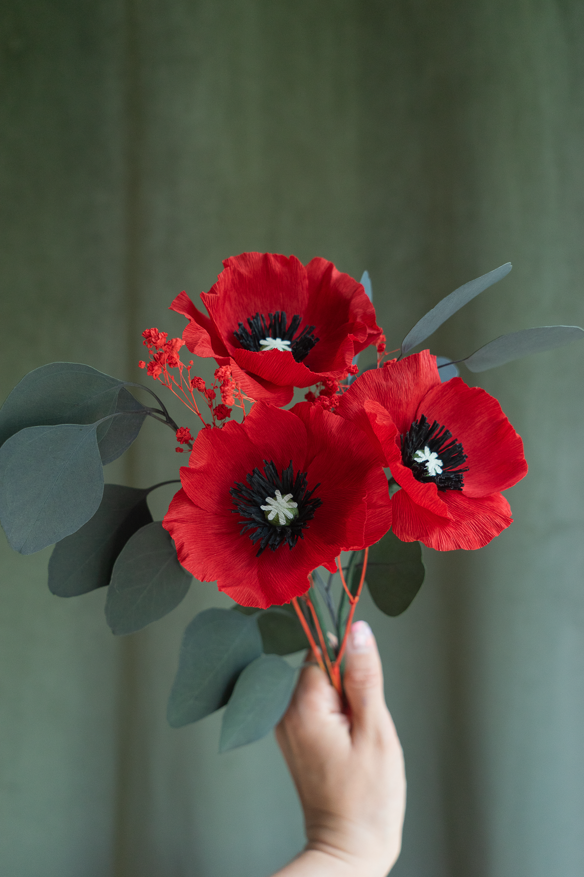Bouquet de trois Coquelicots rouges en papier crépon fait main, accompagnés de gypsophile rouge et de feuilles d'eucalyptus stabilisé, tenu à la main face à un fond velour vert, fleurs éternelles artisanales faites main par Maison Tessier.