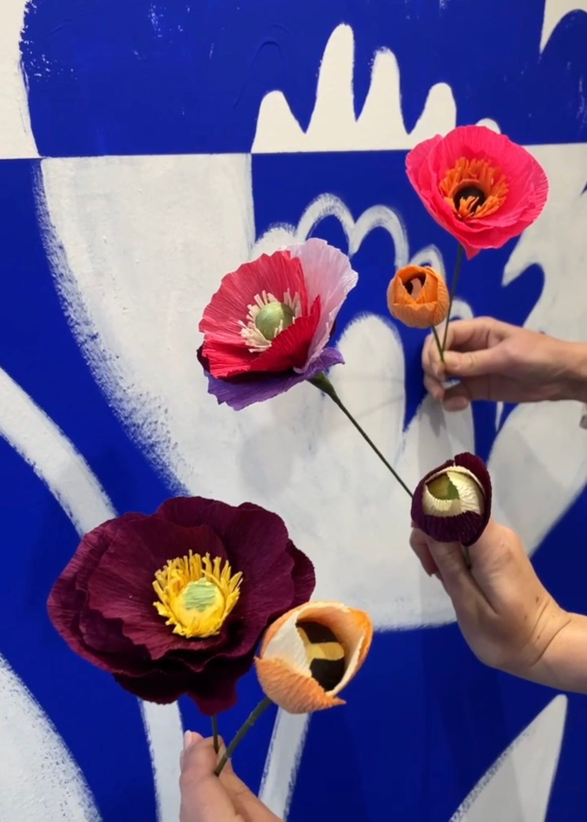 Créations de trois participantes à l’atelier fleurs en papier, trois Pavots de différentes couleurs : un bordeaux, un fusil et un violet, rose clair et fushia, mais aussi trois tulipes : une orange clair, une orange clair et jaune pâle et une bordeaux et jaune pâle, tenus à la main face à un fond bleu et blanc, atelier créatif pour EVJF, team building, animé par Maison Tessier.