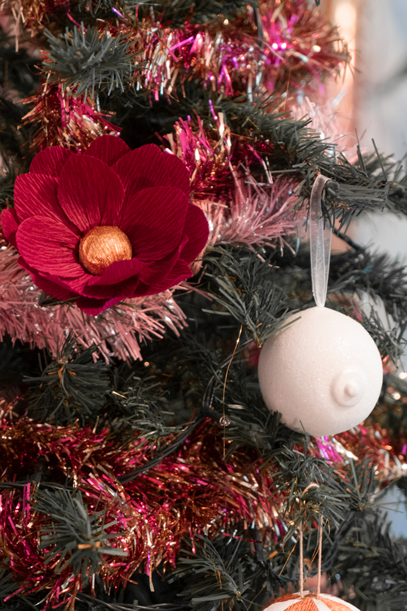 Le Saint des Seins en forme de sein avec paillettes blanche et le téton blanc nacré accroché sur une branche de sapin de Noël décoré de guirlandes, avec une fleur en papier bordeaux coeur or de Maison Tessier.