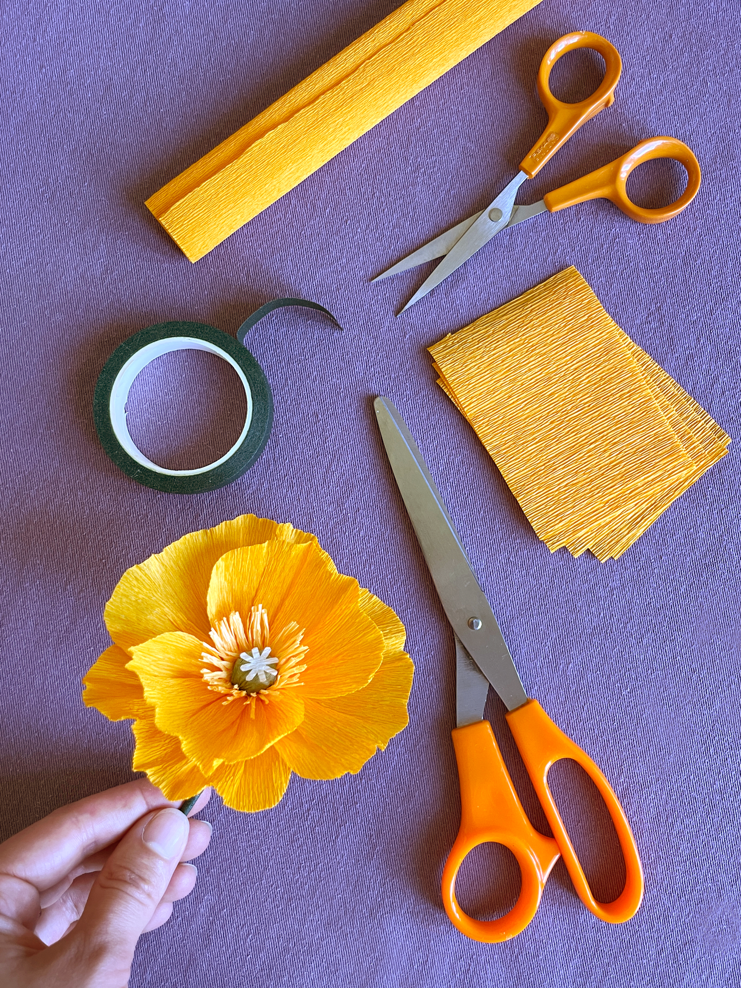 Vue de dessus du matériel nécessaire à la création de fleurs en papier crépon lors d'un atelier, team building, EVJF, Maison Tessier : deux paires de ciseaux orange, des bandes de papier crépon jaune, des pétales prédécoupés et un rouleau de ruban vert, avec le résultat un Pavot jaune terminé, tenu à la main, disposé sur une surface mauve.