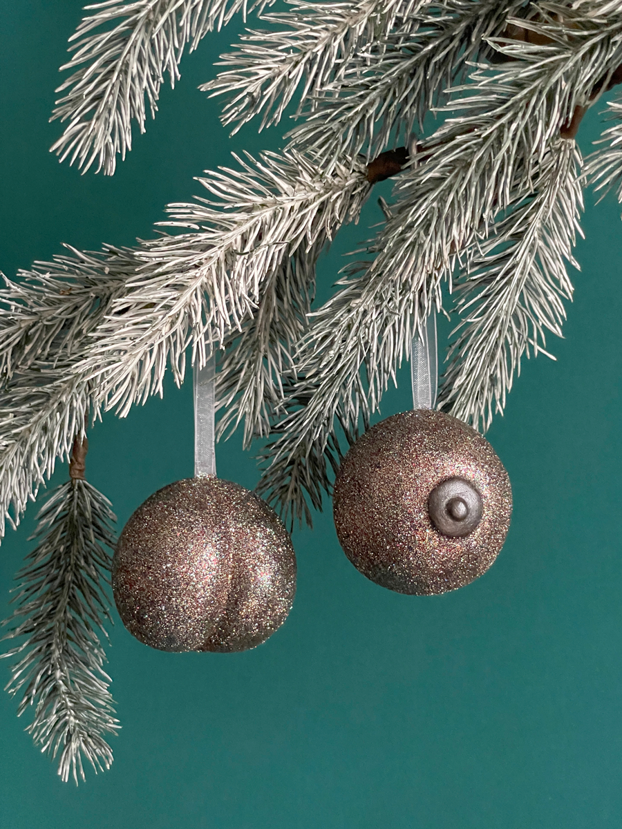 Paire de boules de Noël Maison Tessier, avec le Boule de Noël en forme fesses avec des paillettes bronze et du Saint des Seins avec des paillettes bronze et le téton bronze accrochés sur une branche de sapin, sur fond vert émeraude.