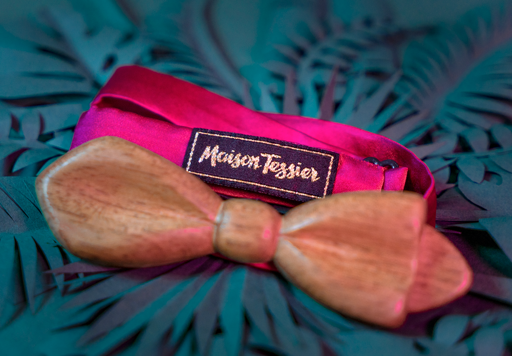 Tour de cou fushia avec un noeud papillon en bois Maison Tessier sur fond de feuilles tropicales en papier, vue de 3/4