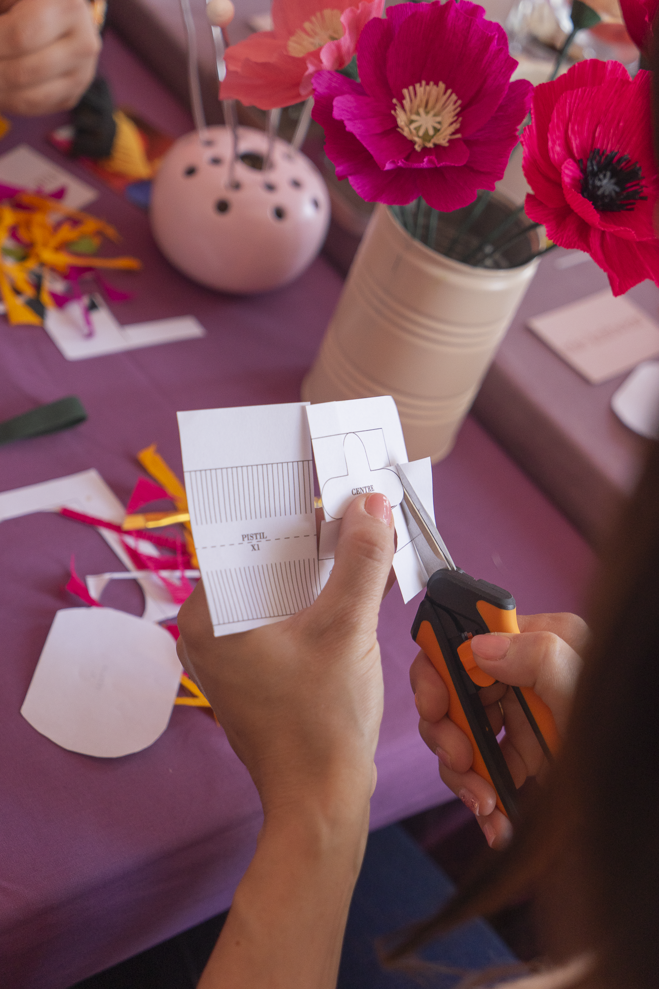 Gros plan de mains découpant un patron de pistil et de coeur d’une fleur aux ciseaux lors d’un atelier fleurs en papier, team building, EVJF, animé par Maison Tessier, avec des morceaux de papier crépon découpés en arrière-plan sur une table avec une nappe violette.