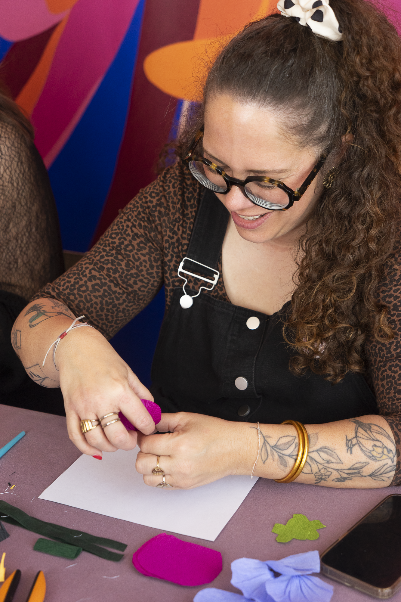 Femme souriante concentrée sur la fabrication d'une fleur en papier crépon fuchsia lors d'un atelier Maison Tessier devant un mur coloré, atelier créatif fleurs en papier, team building, EVJF, animé par Maison Tessier.