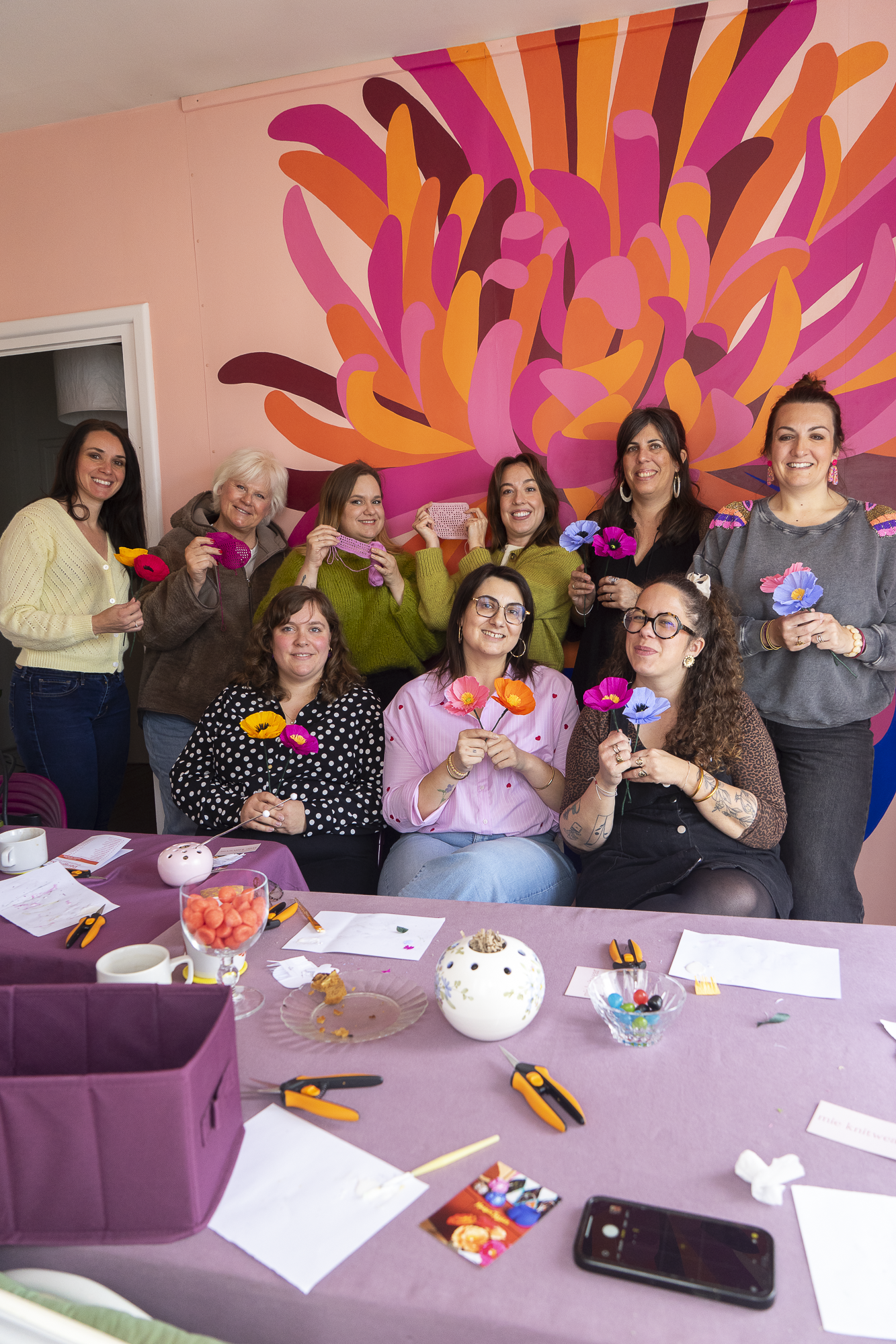 Groupe de neuf participantes souriantes présentant leurs Pavots en papier crépon colorés lors d'un atelier Maison Tessier devant un mur fleuri rose et orange à La Rochelle,  atelier créatif fleurs en papier, team building, EVJF, animé par Maison Tessier.