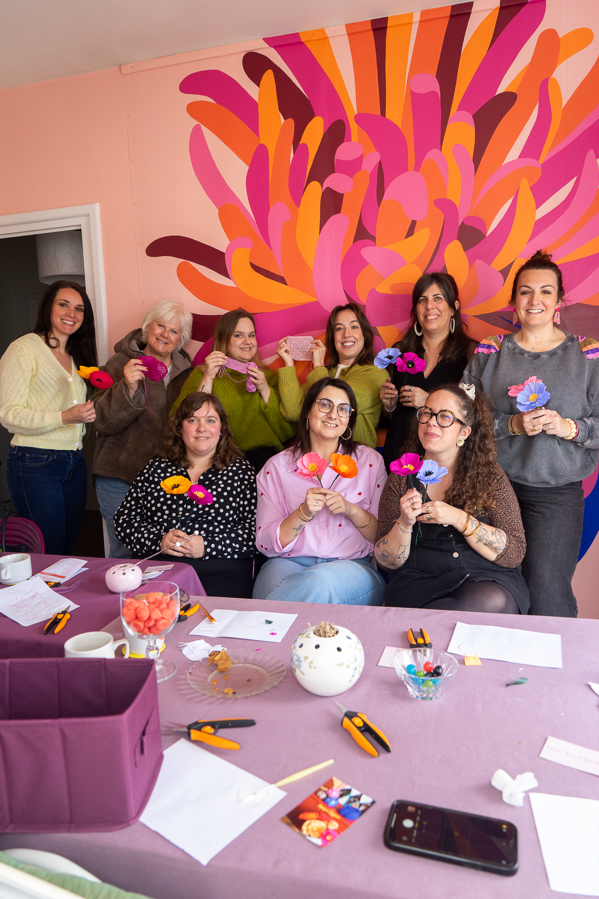 Groupe de neuf participantes souriantes présentant leurs Pavots en papier crépon colorés lors d'un atelier Maison Tessier devant un mur fleuri rose et orange à La Rochelle,  atelier créatif fleurs en papier, team building, EVJF, animé par Maison Tessier.