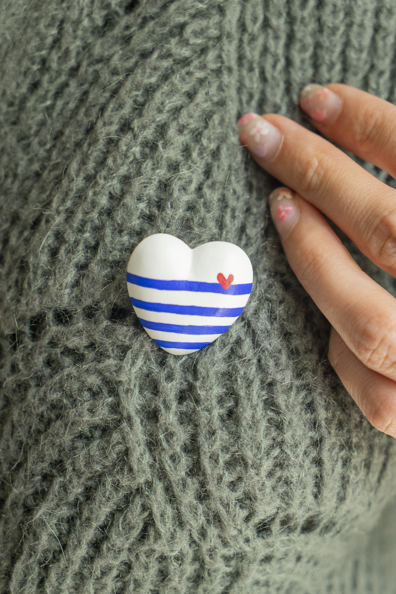 Broche Mini Coeur Marinière Cobalt en résine écologique peinte en avec des lignes de bleu Klein avec un petit coeur rouge en haut à droite, accrochée sur un pull kaki avec une main sur le côté, bijou original fabriqué avec amour par Maison Tessier.