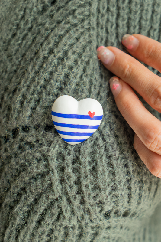 Broche Mini Coeur Marinière Cobalt en résine écologique peinte en avec des lignes de bleu Klein avec un petit coeur rouge en haut à droite, accrochée sur un pull kaki avec une main sur le côté, bijou original fabriqué avec amour par Maison Tessier.