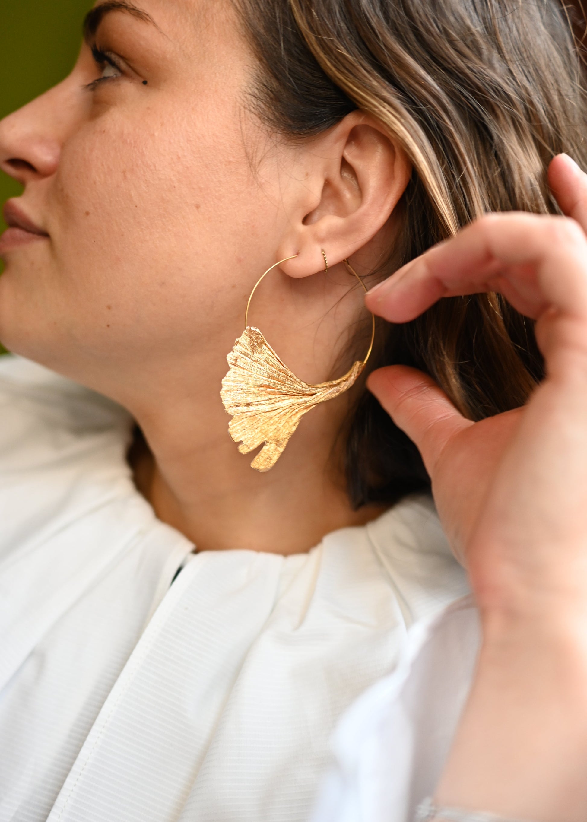 Gros plan de profil sur une femme soulevant ses cheveux pour montrer sa créole Ginkgo dorée en papier sur anneau doré, boucles d'oreilles feuille de ginkgo artisanales faites main par Maison Tessier.
