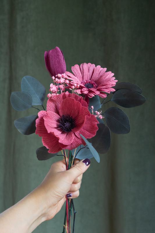 Bouquet de trois fleurs composé d’un Gerbera vieux rose, une Tulipe prun et un Pavot vieux rose, accompagné de gypsophile stabilisé rose et d’eucalyptus séchées, tenus à la main sur un fond vert kaki, fleurs artisanales faites main par Maison Tessier 