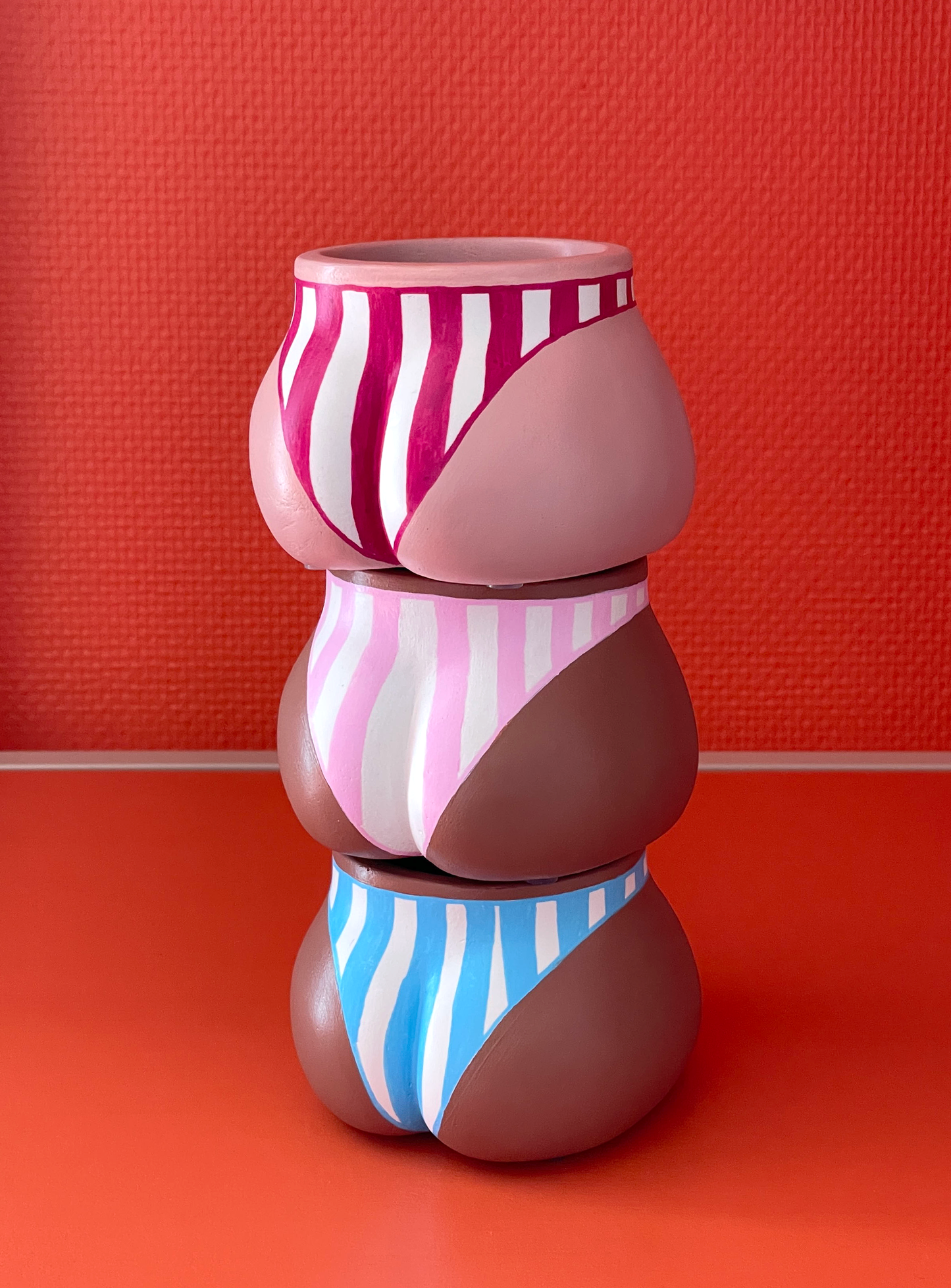 Cache-pots Fessier côté fesses en résine écologique fait main présentés empilés avec les trois coloris : crème avec maillot de bain à rayures verticales grenat, café avec maillot de bain à rayures roses et café avec maillot de bain à rayures bleues, sur un fond orange corail, objet déco original corps féminin fait main par Maison Tessier.