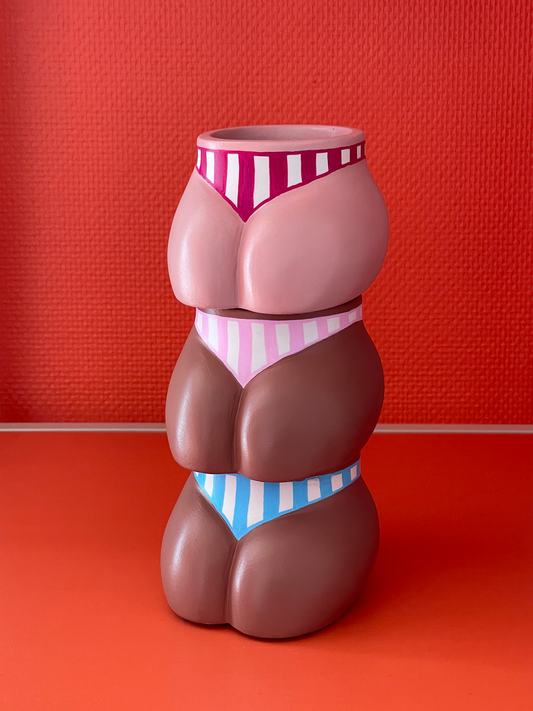 Cache-pots Fessier côté pubis en résine écologique fait main présentés empilés avec les trois coloris : crème avec maillot de bain à rayures verticales grenat, café avec maillot de bain à rayures roses et café avec maillot de bain à rayures bleues, sur un fond orange, objet déco original corps féminin fait main par Maison Tessier.