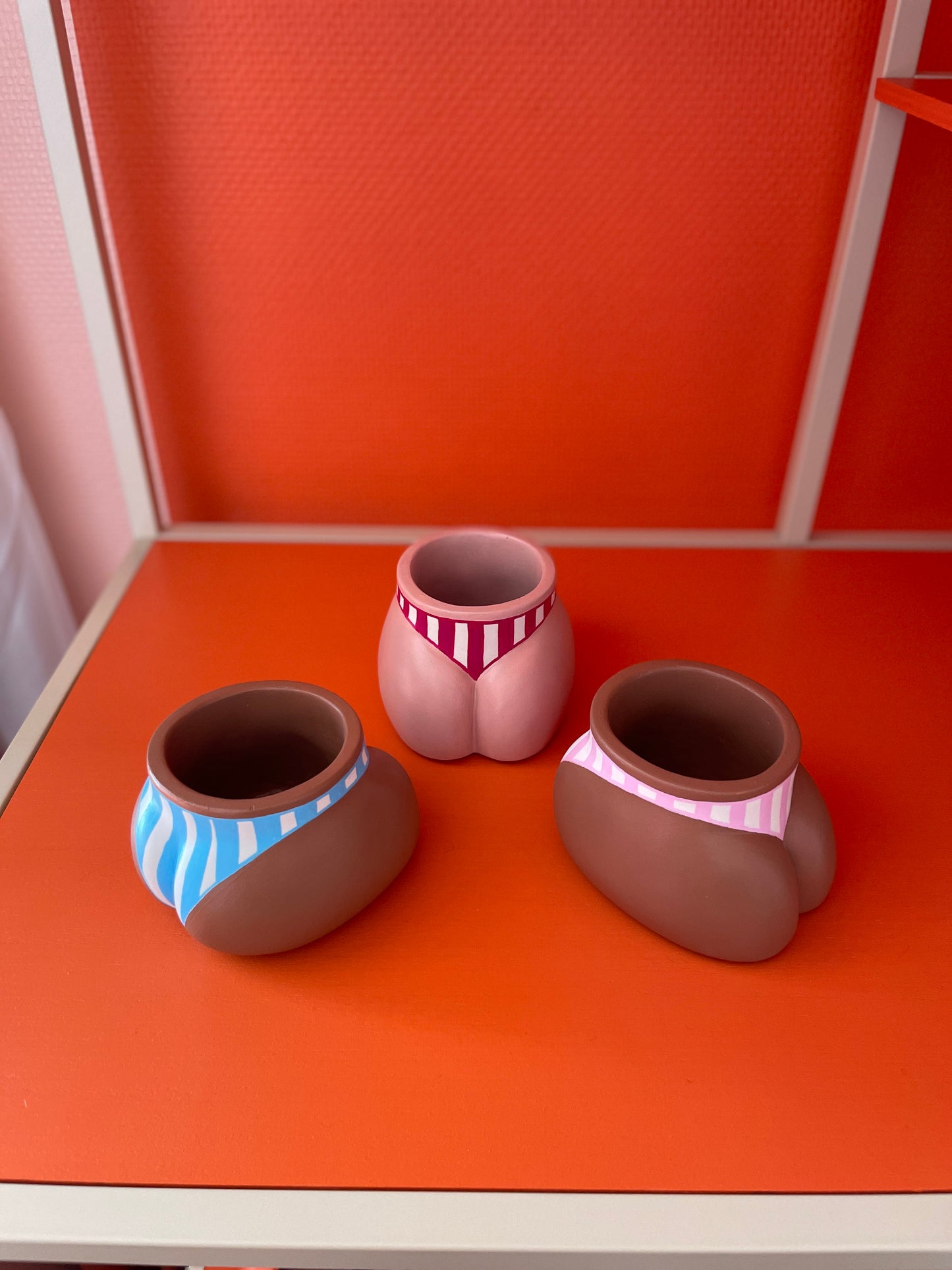 Les trois cache-pots Fessier de la collection Cabanon présentés ensemble sur un fond orange : côté pubis coloris crème avec maillot de bain à rayures grenat au centre, côté fesses à gauche, coloris café avec maillot de bain à rayures bleues et à droite, café avec maillot de bain à rayures roses clair, objet déco original corps féminin fait main en résine écologique (Jesmonite) par Maison Tessier.