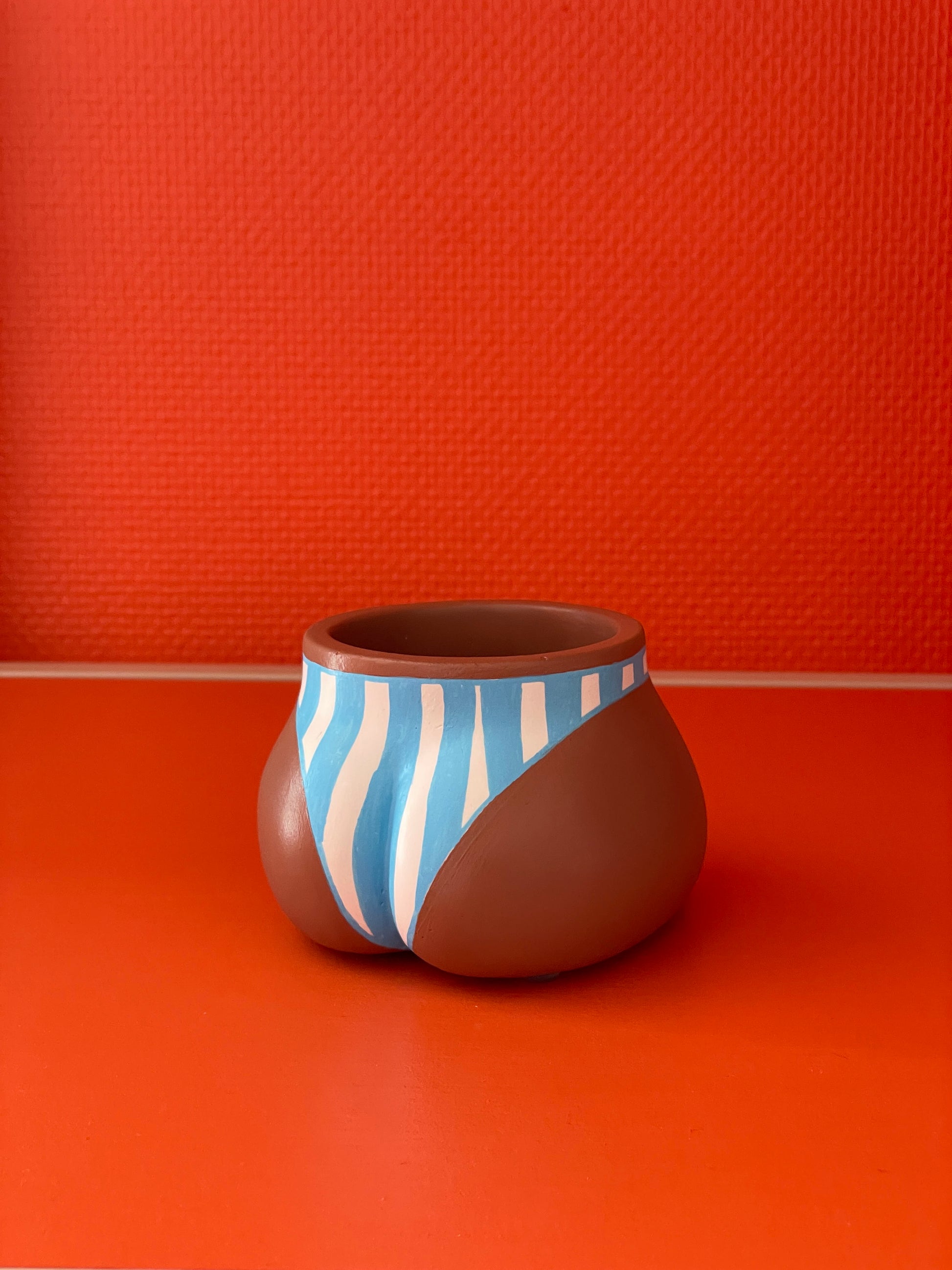 Cache-pot Fessier, côté fesses, de coloris café avec un maillot de bain à larges rayures verticales bleues et blanches sur un fond orange, objet déco original corps féminin fait main en résine écologique (Jesmonite) par Maison Tessier.