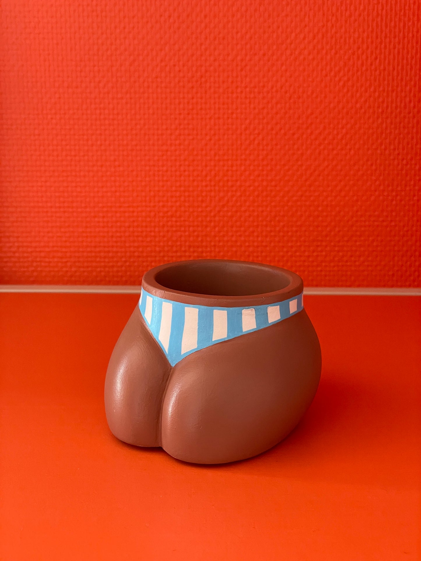 Cache-pot Fessier, côté pubis, de coloris café avec un maillot de bain à larges rayures verticales bleues et blanches sur un fond orange, objet déco original corps féminin fait main en résine écologique (Jesmonite) par Maison Tessier.
