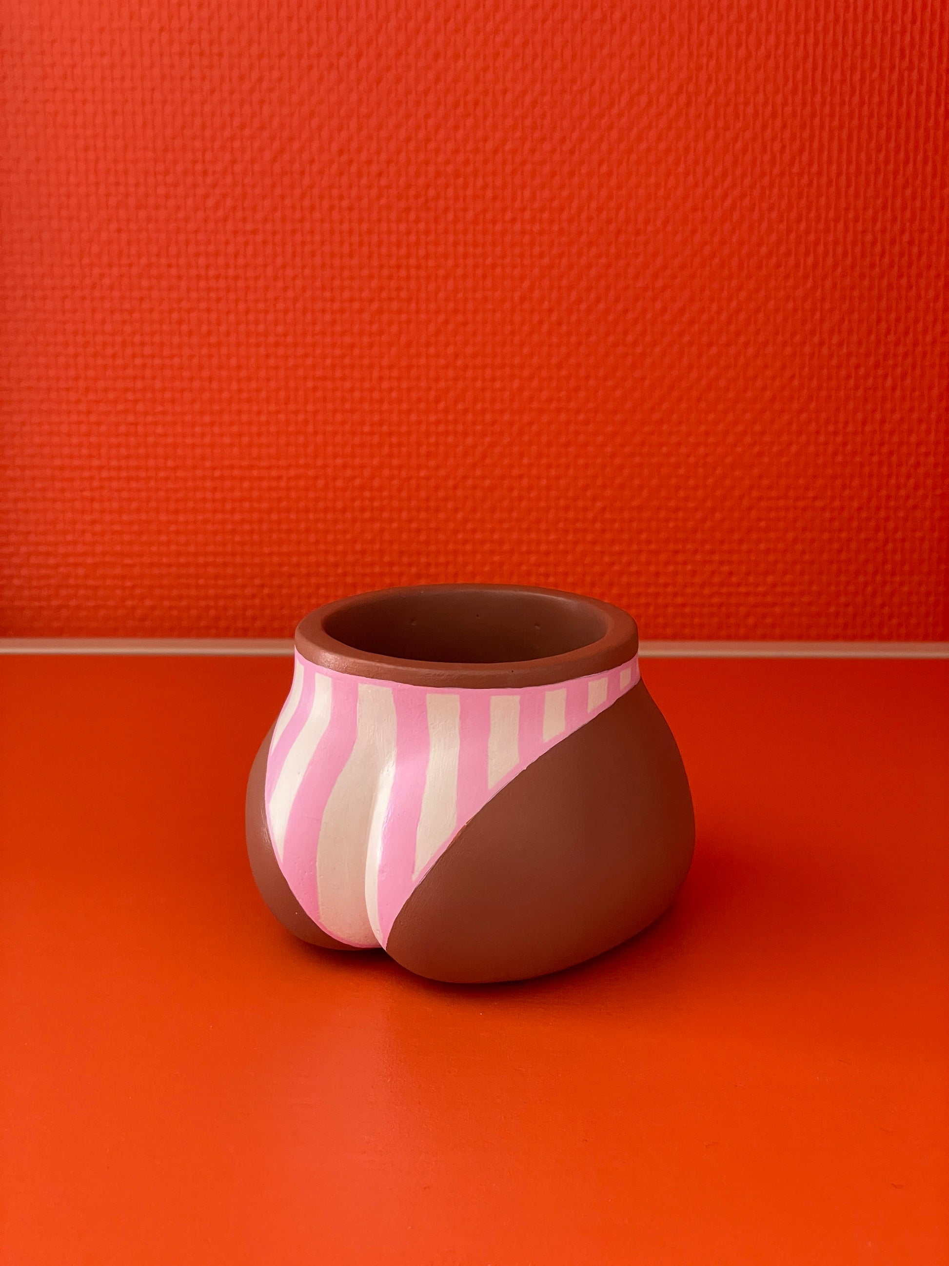 Cache-pot Fessier côté fesses coloris café avec un maillot de bain à rayures verticales roses et crème sur un fond orange, objet déco original corps féminin fait main en résine écologique (Jesmonite) par Maison Tessier.
