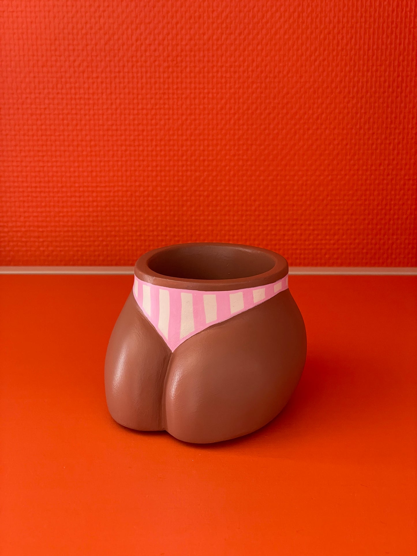 Cache-pot Fessier côté pubis coloris café avec un maillot de bain à rayures verticales roses et crème sur un fond orange, objet déco original corps féminin fait main en résine écologique (Jesmonite) par Maison Tessier.
