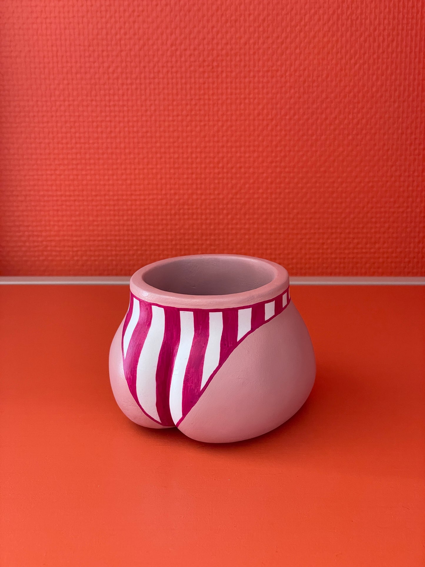 Vue de trois quarts côté pubis du cache-pot Fessier coloris crème avec un maillot de bain à larges rayures verticales grenat et blanc sur un fond orange, objet déco original corps féminin fait main en résine écologique (Jesmonite) par Maison Tessier.