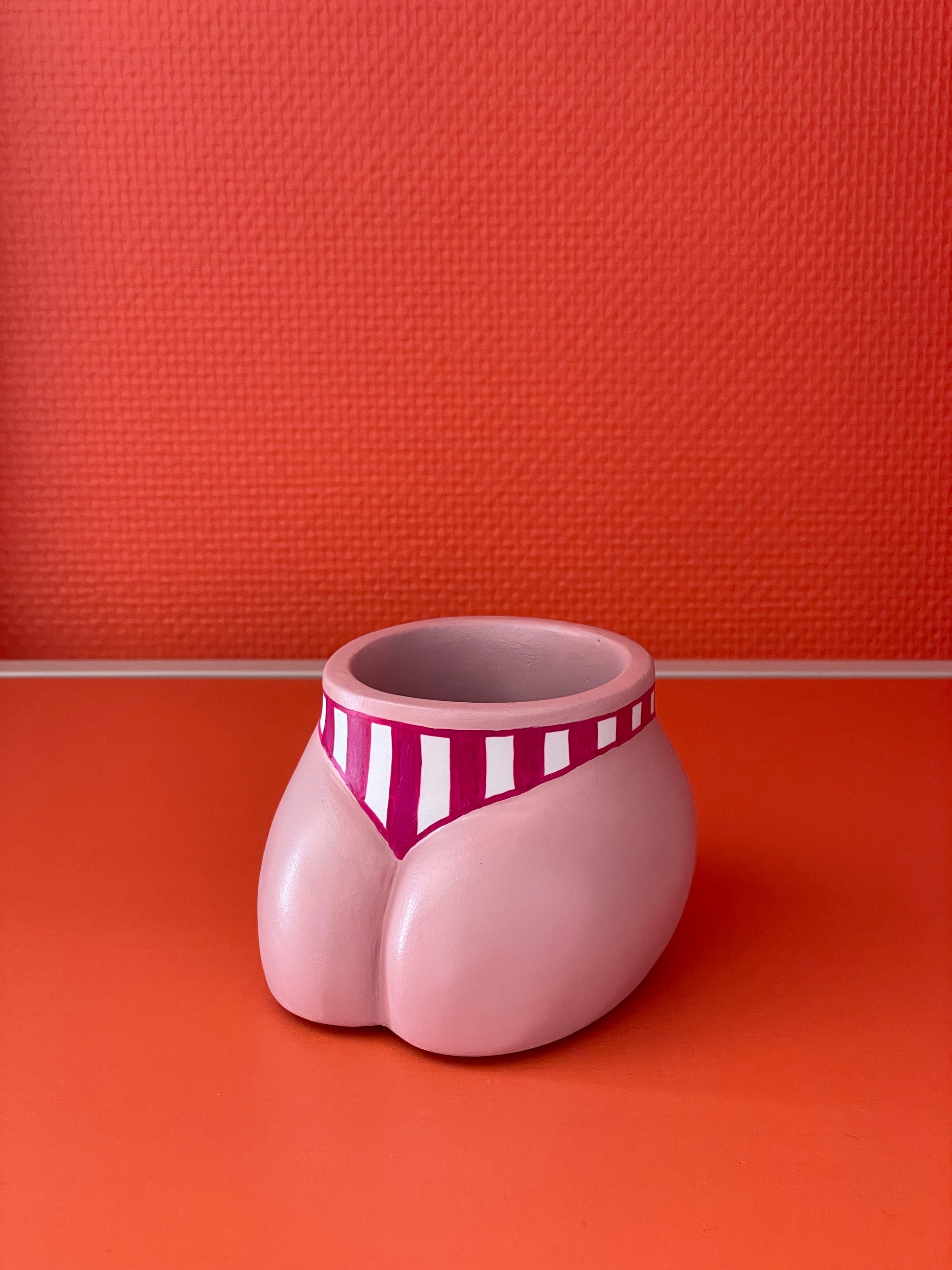 Cache-pot Fessier côté pubis coloris crème avec un maillot de bain à larges rayures verticales grenat et blanc sur un fond orange, objet déco original corps féminin fait main en résine écologique (Jesmonite) par Maison Tessier.