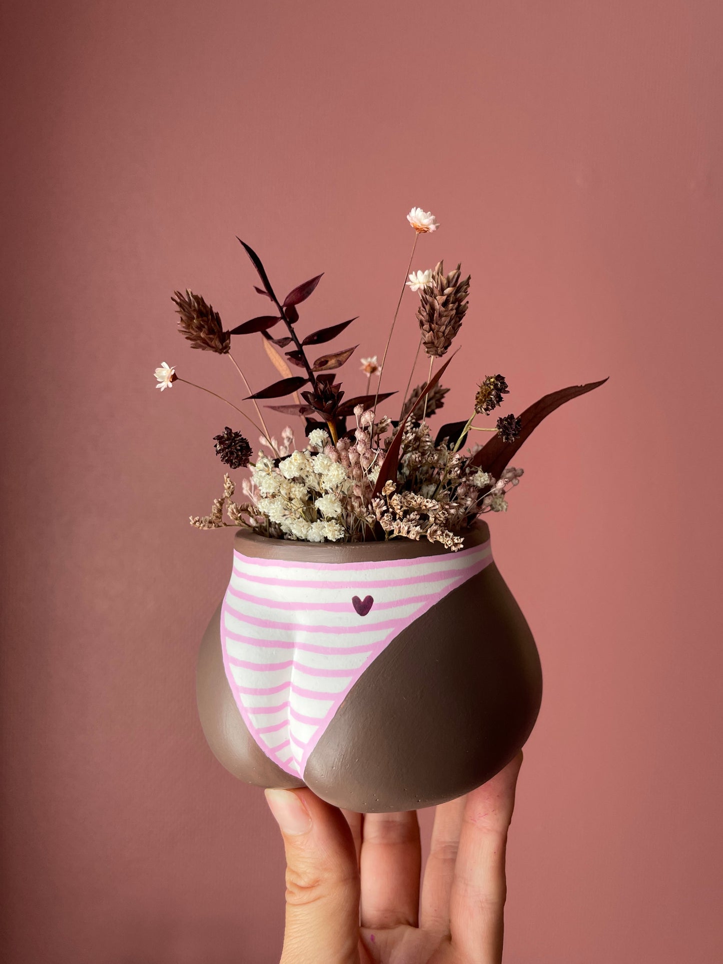 Main tenant un mini cache-pot Fessier en résine écologique, côté fesses, couleur peau noire avec une culotte rayures roses et blanches, avec un petit cœur, garni d'un bouquet de fleurs séchées, gypsophiles, glixias, phalaris, hortensias colorées brunes et blanches sur fond rose, objet déco floral original fait main par Maison Tessier