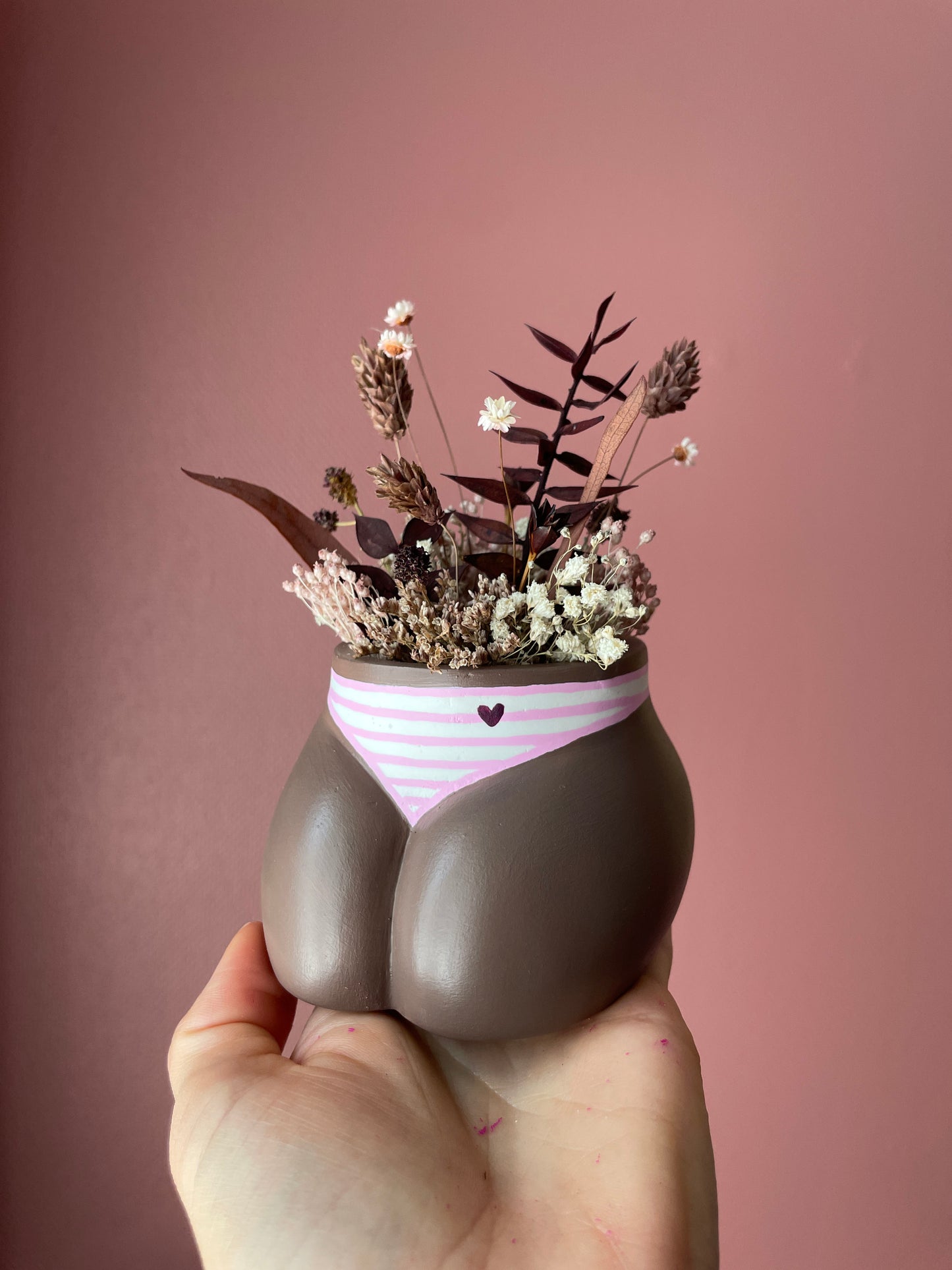 Main tenant un mini cache-pot Fessier en résine écologique, côté pubis, couleur peau noire avec une culotte rayures roses et blanches, avec un petit cœur, garni d'un bouquet de fleurs séchées, gypsophiles, glixias, phalaris, hortensias colorées brunes et blanches sur fond rose, objet déco floral original fait main par Maison Tessier