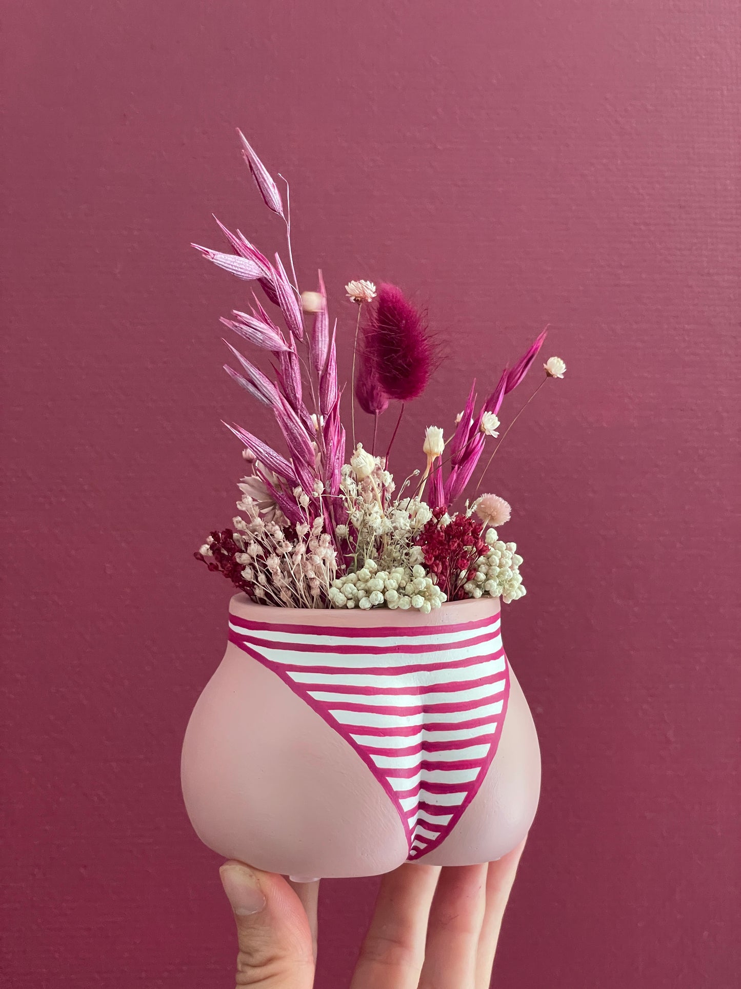 Main tenant un mini cache-pot Fessier, côté fesses, couleur peau blanche en résine écologique avec une culotte rayures fuchsia et blanc garni d'un bouquet de fleurs séchées lagures, avoine, gypsophiles, glixias, phalaris, hortensias, fleurs de riz, colorés fuchsia et blanches sur un fond bordeaux, objet déco floral original fait main par Maison Tessier
