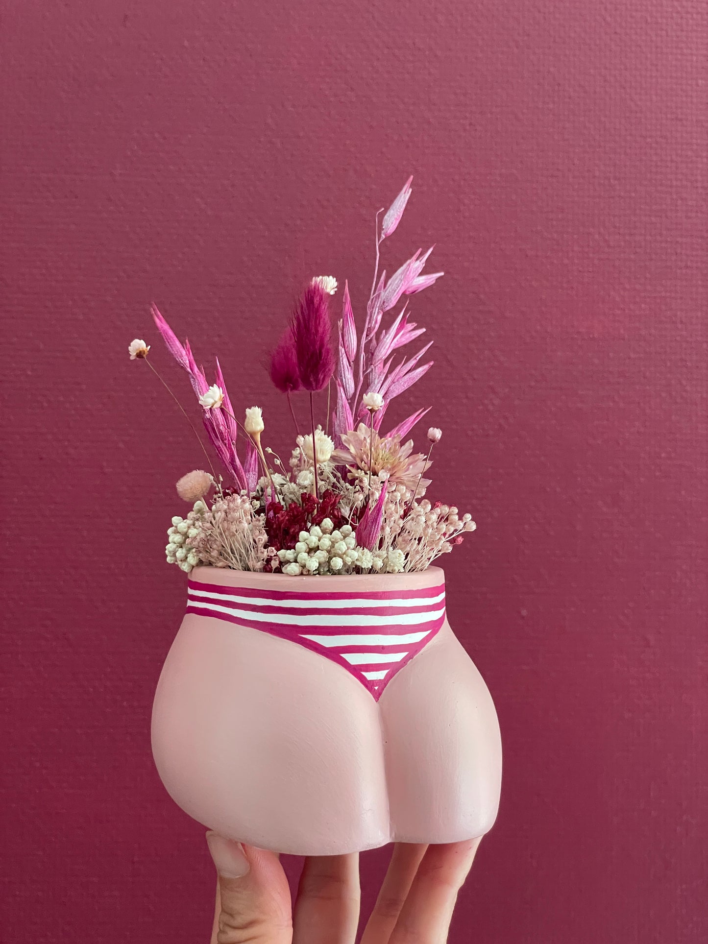 Main tenant un mini cache-pot Fessier, côté pubis, couleur peau blanche en résine écologique avec une culotte rayures fuchsia et blanc garni d'un bouquet de fleurs séchées lagures, avoine, gypsophiles, glixias, phalaris, hortensias, fleurs de riz, colorés fuchsia et blanches sur un fond bordeaux, objet déco floral original fait main par Maison Tessier