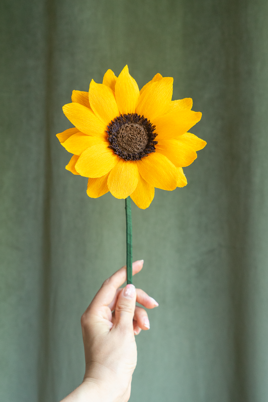 Tournesol en papier crépon jaune et cœur brun tenu à la main sur fond vert kaki, fleur artisanale faite main par Maison Tessier
