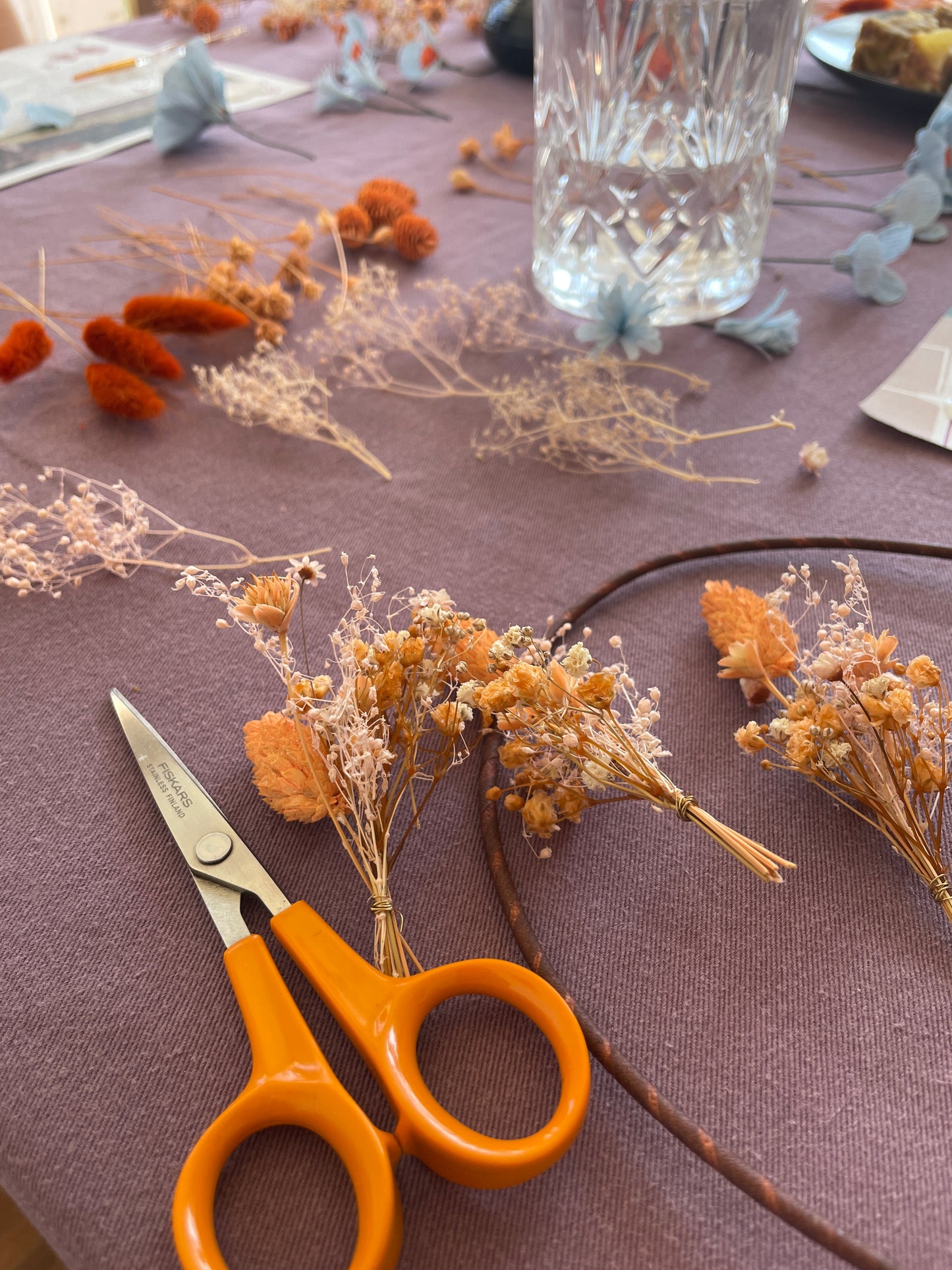 Mini bouquets de fleurs séchées disposés sur la table avec un ciseau pour pouvoir assembler sa couronne de fleurs, en arrière-plan un verre d’eau, atelier créatif pour EVJF, team building, animé par Maison Tessier.