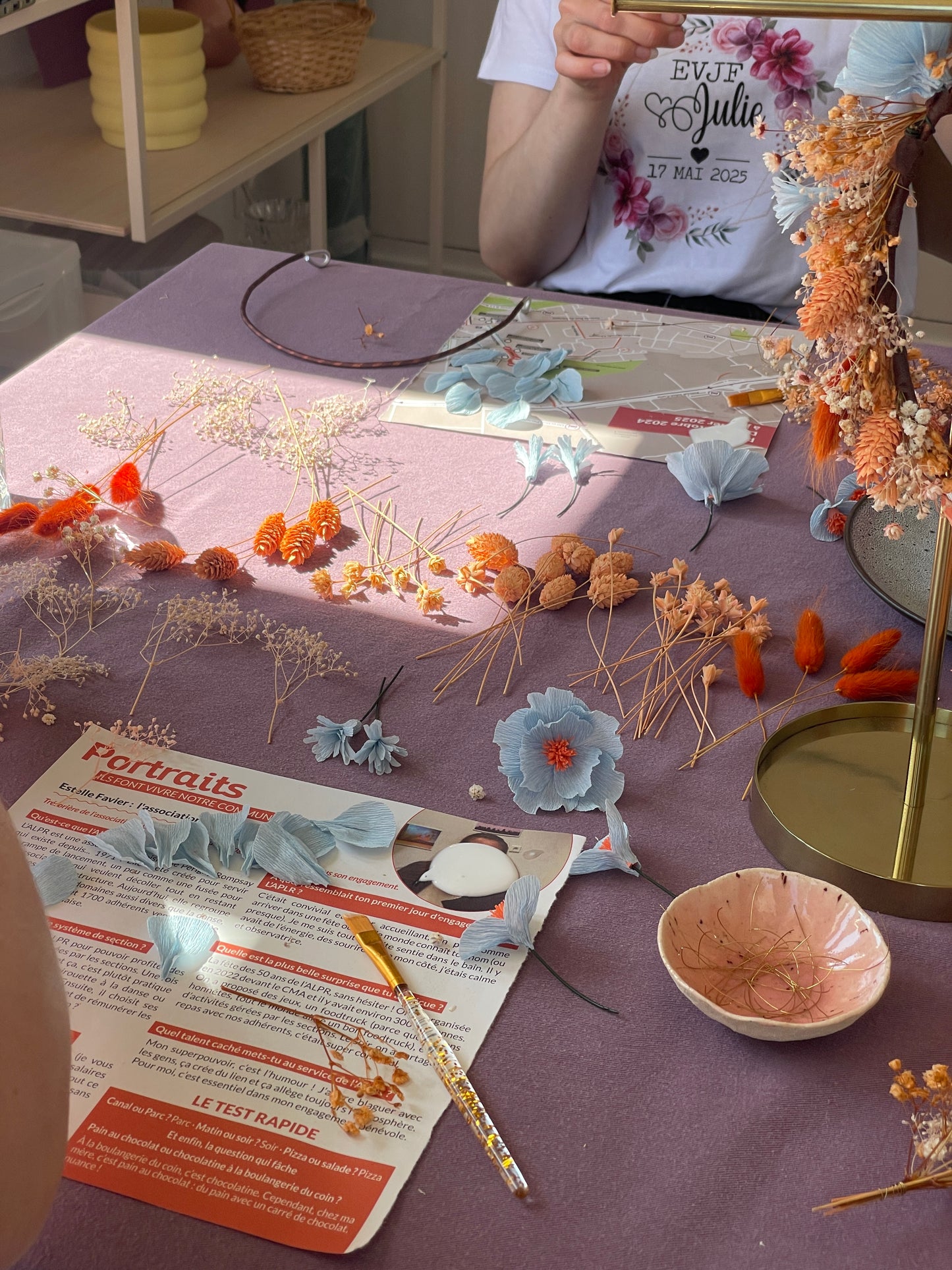 Table d'atelier EVJF à La Rochelle recouverte d'une nappe mauve avec des fleurs en papier crépon bleu pâle à pistils orange, des fleurs séchées dorées et orangées classées par variété, un cerceau en métal et des outils de création, participante portant un t-shirt personnalisé EVJF Julie 17 mai 2025 visible en arrière-plan, atelier créatif, team building, animé par Maison Tessier.