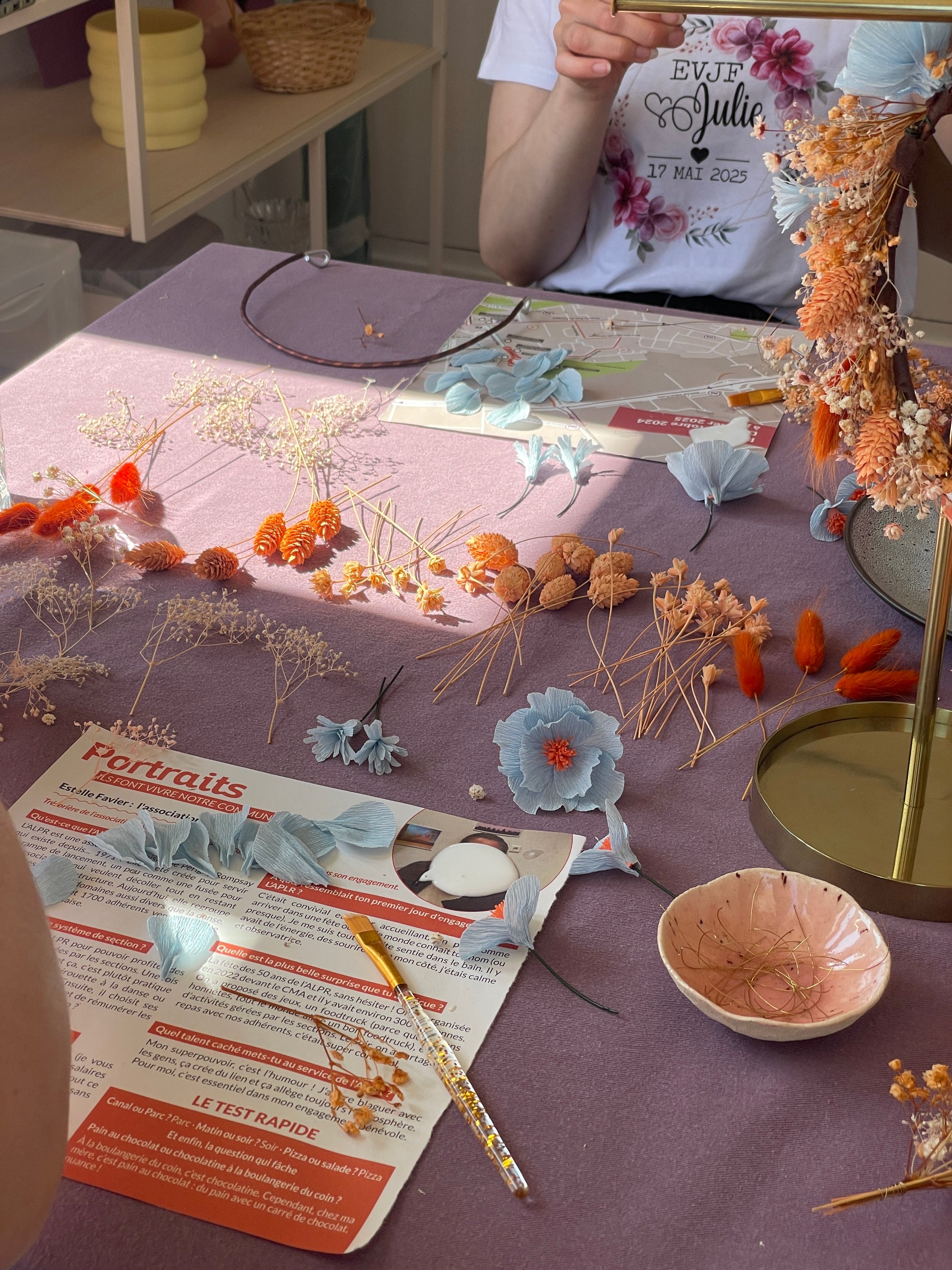 Table d'atelier EVJF à La Rochelle recouverte d'une nappe mauve avec des fleurs en papier crépon bleu pâle à pistils orange, des fleurs séchées dorées et orangées classées par variété, un cerceau en métal et des outils de création, participante portant un t-shirt personnalisé EVJF Julie 17 mai 2025 visible en arrière-plan, atelier créatif, team building, animé par Maison Tessier.
