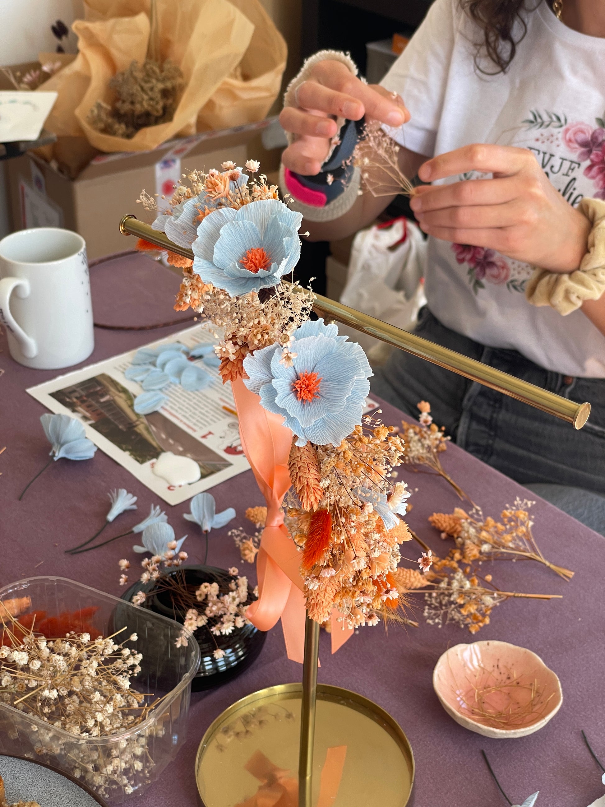 Couronne fleurie en exemple lors d'un atelier EVJF Maison Tessier à La Rochelle, composée d'un cerceau rond décoré de trois fleurs bleu pâle à pistils orange en papier crépon, de fleurs séchées dorées et orangées et d'un ruban satiné pêche, participante en t-shirt EVJF assemblant les fleurs séchées en arrière-plan, atelier créatif pour un EVJF, team building, animé par Maison Tessier.