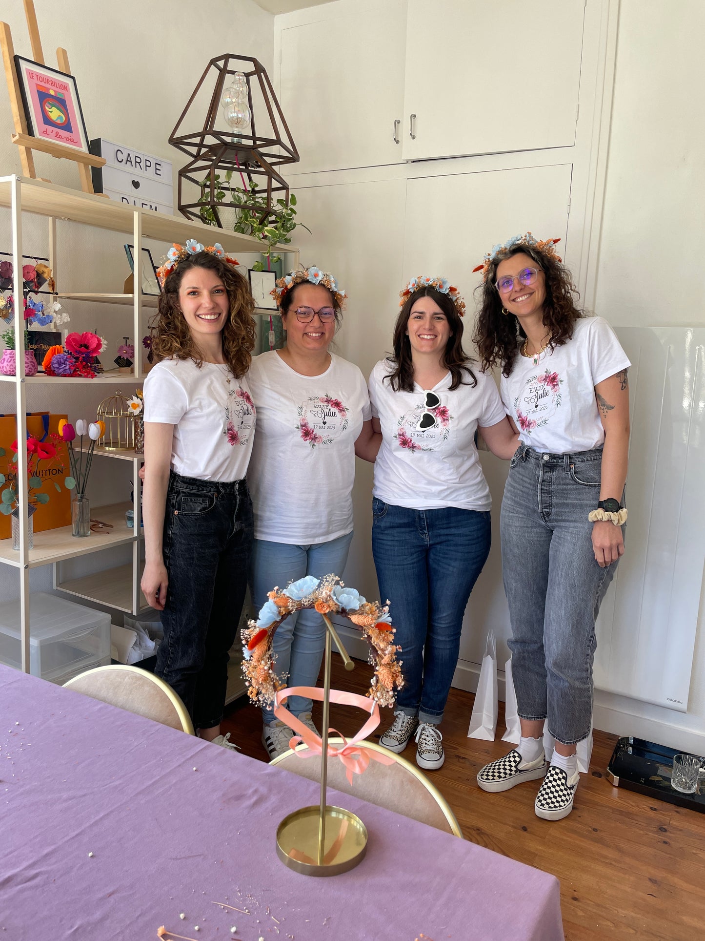 Groupe de 4 participantes avec le sourire portant des tee-shirts EVJF et les couronnes de fleurs bleu et orange qu’elles viennent créer pendant l’atelier, avec devant l’exemple qu’elles avaient et en arrière plan les étagères avec des créations, ateliers EVJF pour se détendre, team building, animé par Maison Tessier.