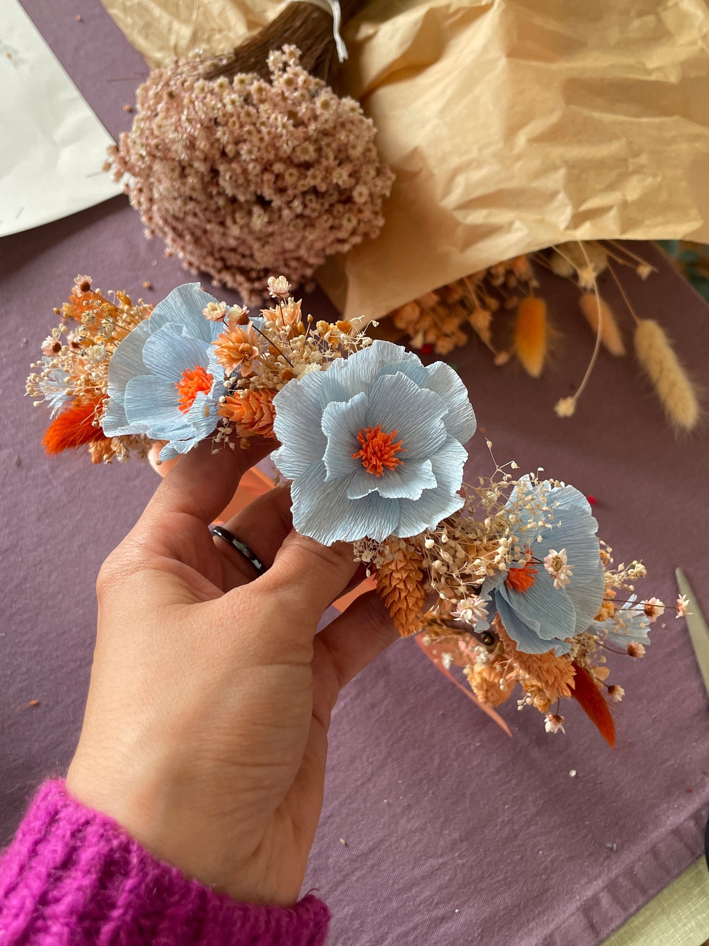 Couronne de fleurs réalisée pendant l’atelier avec des fleurs en papier bleu coeur orange et des fleurs séchées, tenue par une main avec en arrière-plan les bouquets de fleurs séchées, atelier créatif privatisé, team building, EVJF, animé par Maison Tessier.