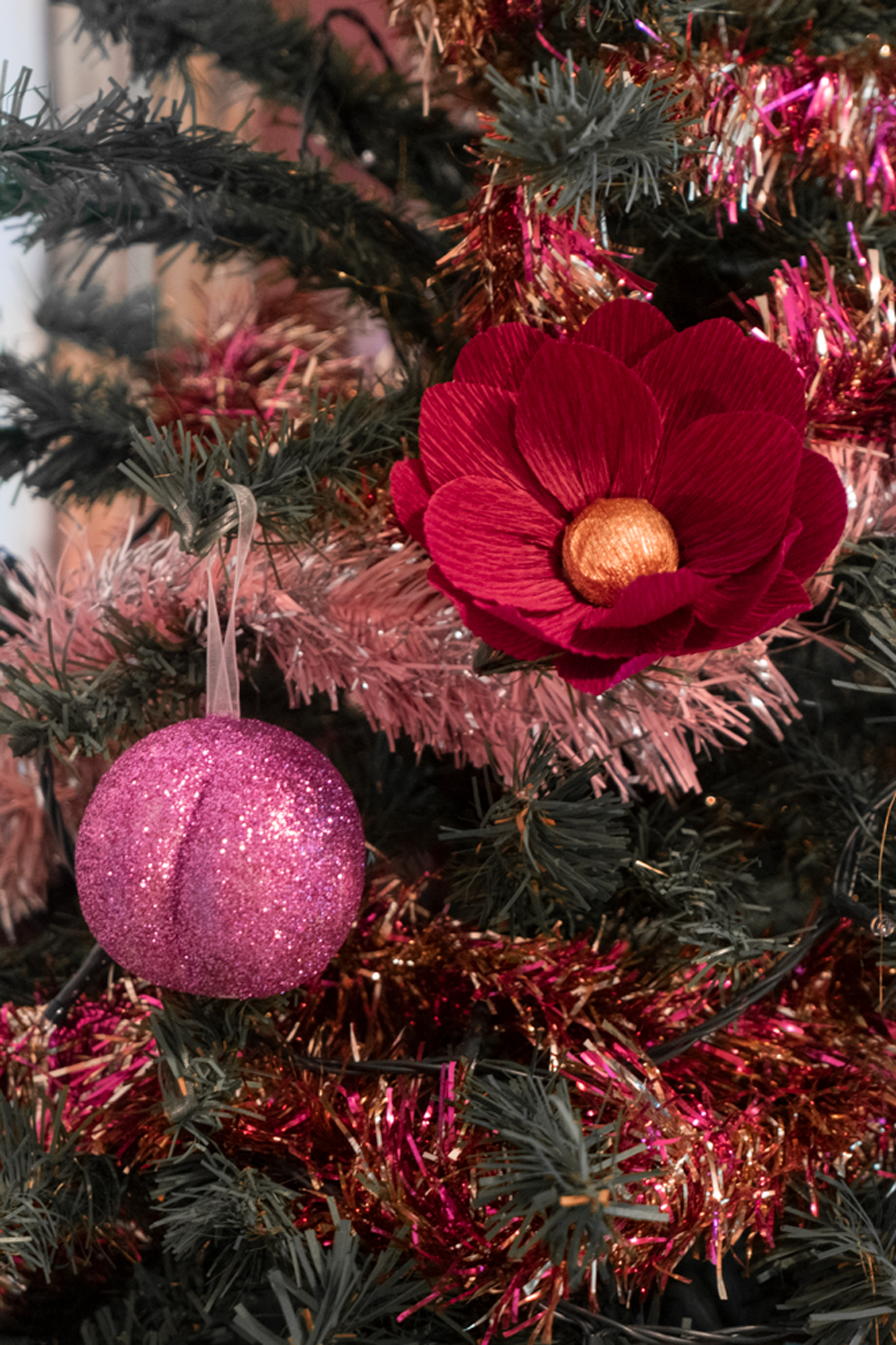 Le Boule de Noël en forme de  fesses avec des paillettes roses accroché sur une branche de sapin de Noël décoré de guirlandes, avec une fleur en papier bordeaux coeur or de Maison Tessier.