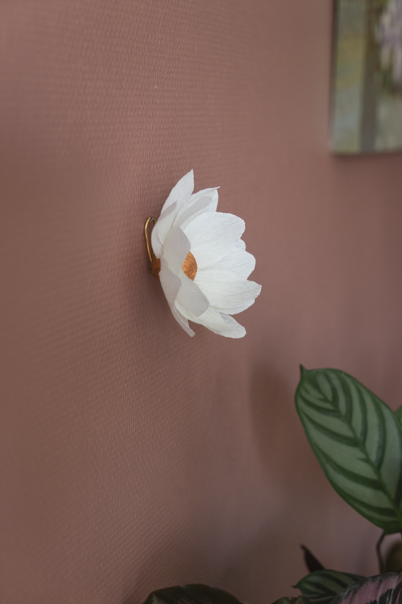 décoration murale florale faite à la main par Maison Tessier, fleur en papier Nymphea, de couleur blanche au coeur cuivrée, accrochée sur un mur rose texturé, accompagné de plantes d'intérieurs, calathea, et d'un tableau, vue de côté pour voir accroche murale avec un clou