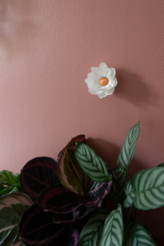 Fleur en papier crépon Nymphea blanche avec le coeur cuivre accroché sur un mur vieux rose avec des plantes calâtes en dessous, une fleur artisanale fabriquée par Maison Tessier