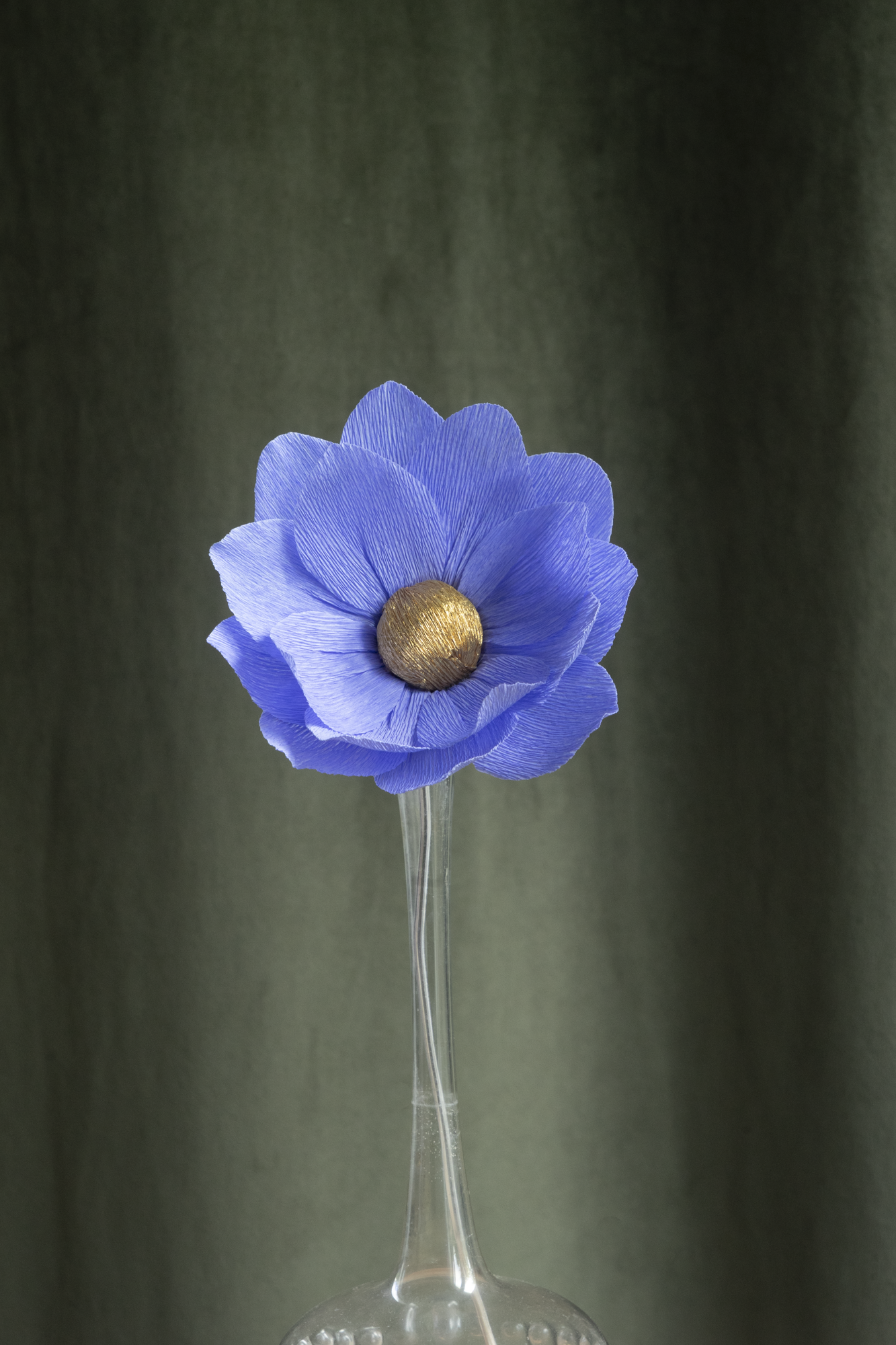 décoration florale faite à la main par Maison Tessier, fleur en papier Nymphea, de couleur bleue au coeur or, présentée dans un vase en verre transparent, sur fond d'un tissus velour vert kaki