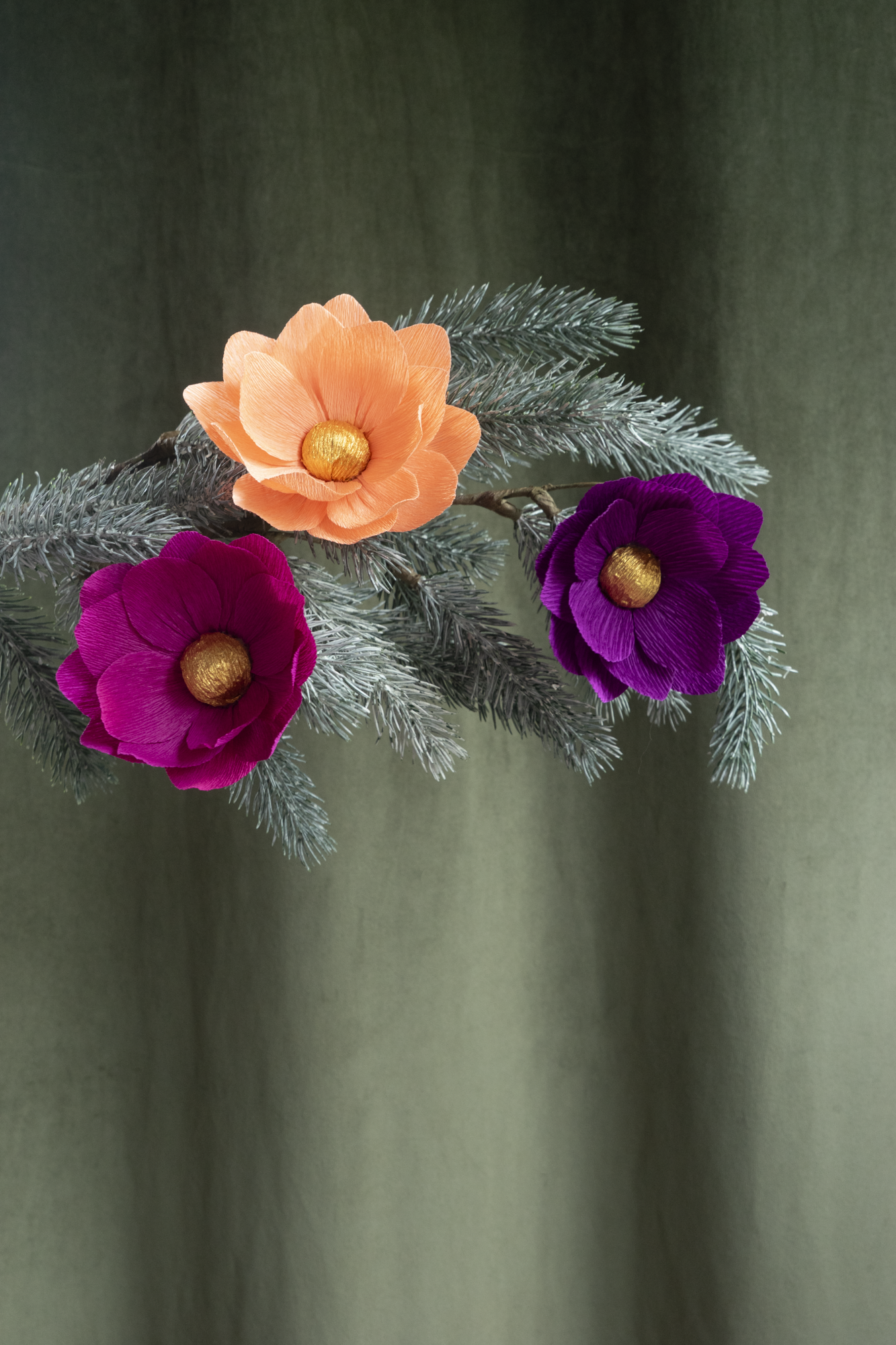 3 fleurs en papier de Maison Tessier , fushia, orange clair et violet sont accrochées sur une branche de sapin de Noel, sur un fond d'un tissus velour kaki 