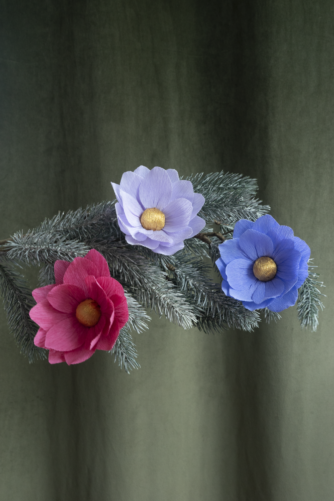 3 fleurs en papier de Maison Tessier , rose, parme et bleue, sont accrochées sur une branche de sapin de Noel, sur un fond d'un tissus velour kaki 