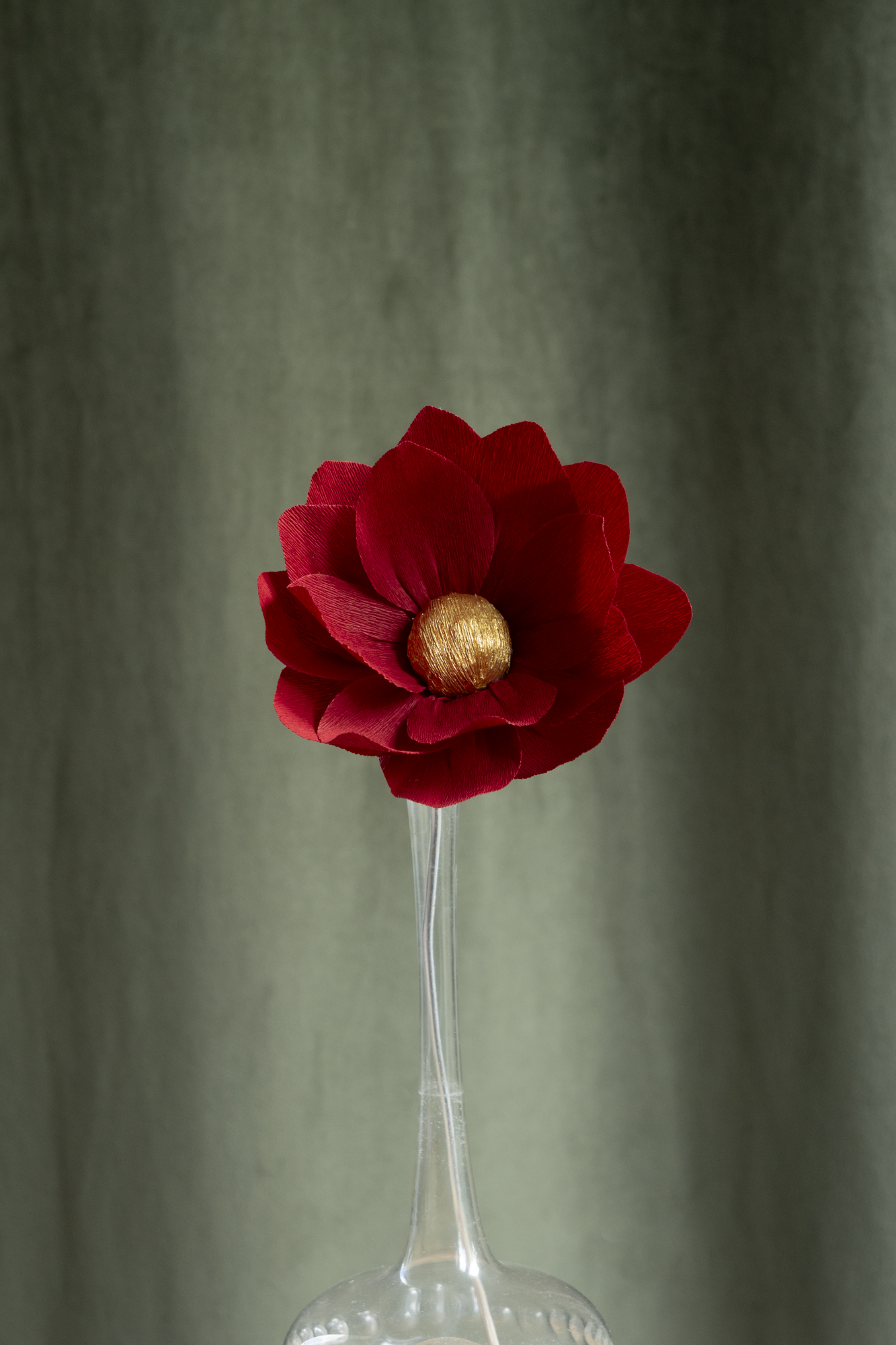 décoration florale faite à la main par Maison Tessier, fleur en papier Nymphea, de couleur rouge foncé au coeur or, présentée dans un vase en verre transparent, sur fond d'un tissus velour vert kaki