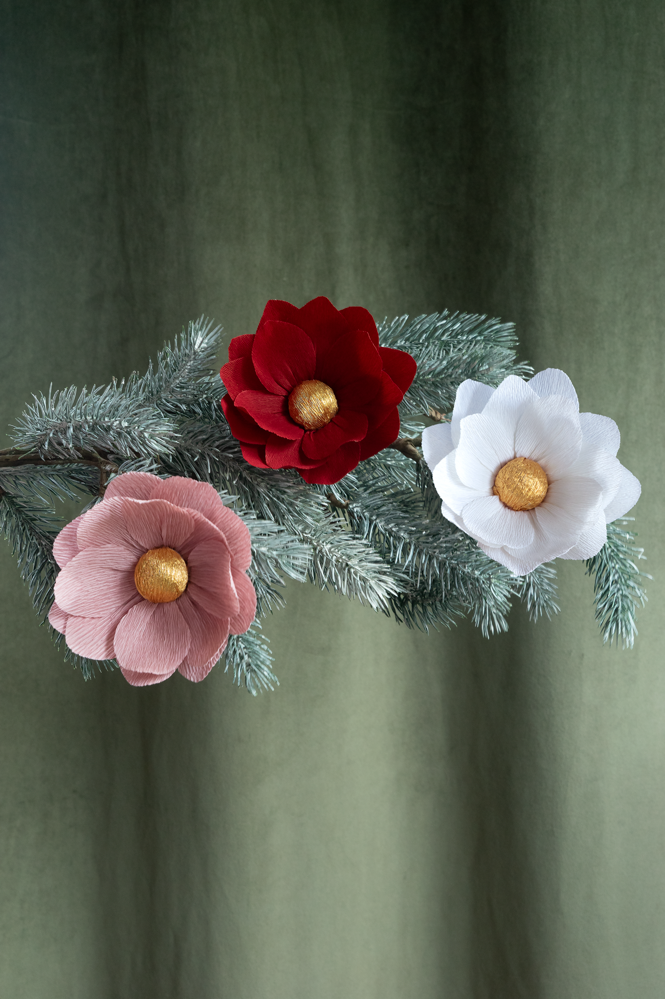3 fleurs en papier de Maison Tessier , vieux rose, rouge foncé et blanche, sont accrochées sur une branche de sapin de Noel, sur un fond d'un tissus velour kaki 