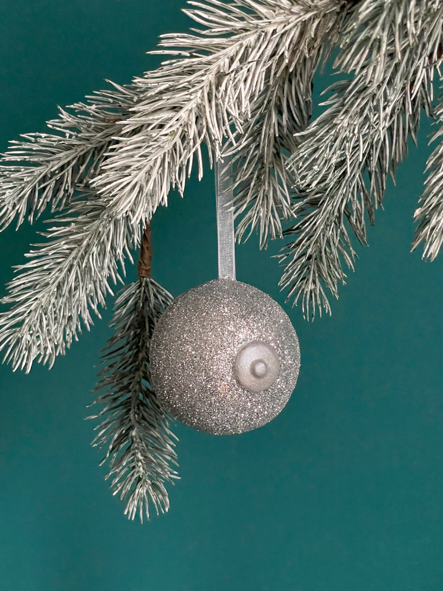 Le Saint des Seins en forme de sein avec paillettes argent et le téton argent accroché sur une branche de sapin, sur fond vert émeraude.