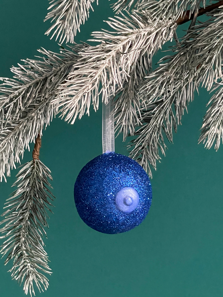 Le Saint des Seins en forme de sein avec paillettes bleues et le téton bleu nacré accroché sur une branche de sapin, sur fond vert émeraude.